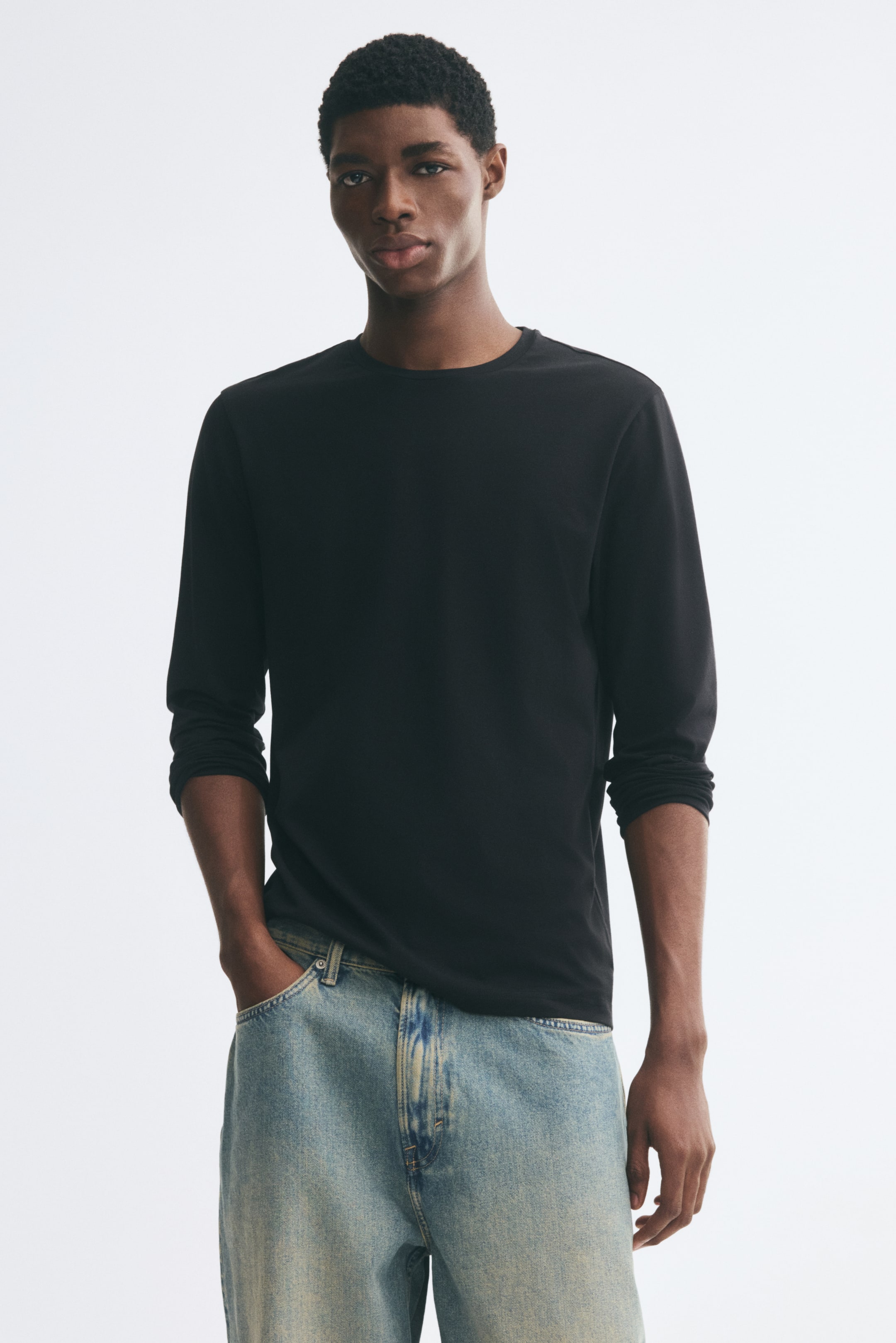 Größeres Bild anzeigen: Ein schwarzes Langarm-T-Shirt mit Rundhalsausschnitt wird vom Model getragen und mit hell gewaschenen Denim-Jeans kombiniert. Das T-Shirt hat eine lockere Passform, die Ärmel sind leicht hochgeschoben, und das Model steht mit einer Hand in der Gesäßtasche der Jeans.