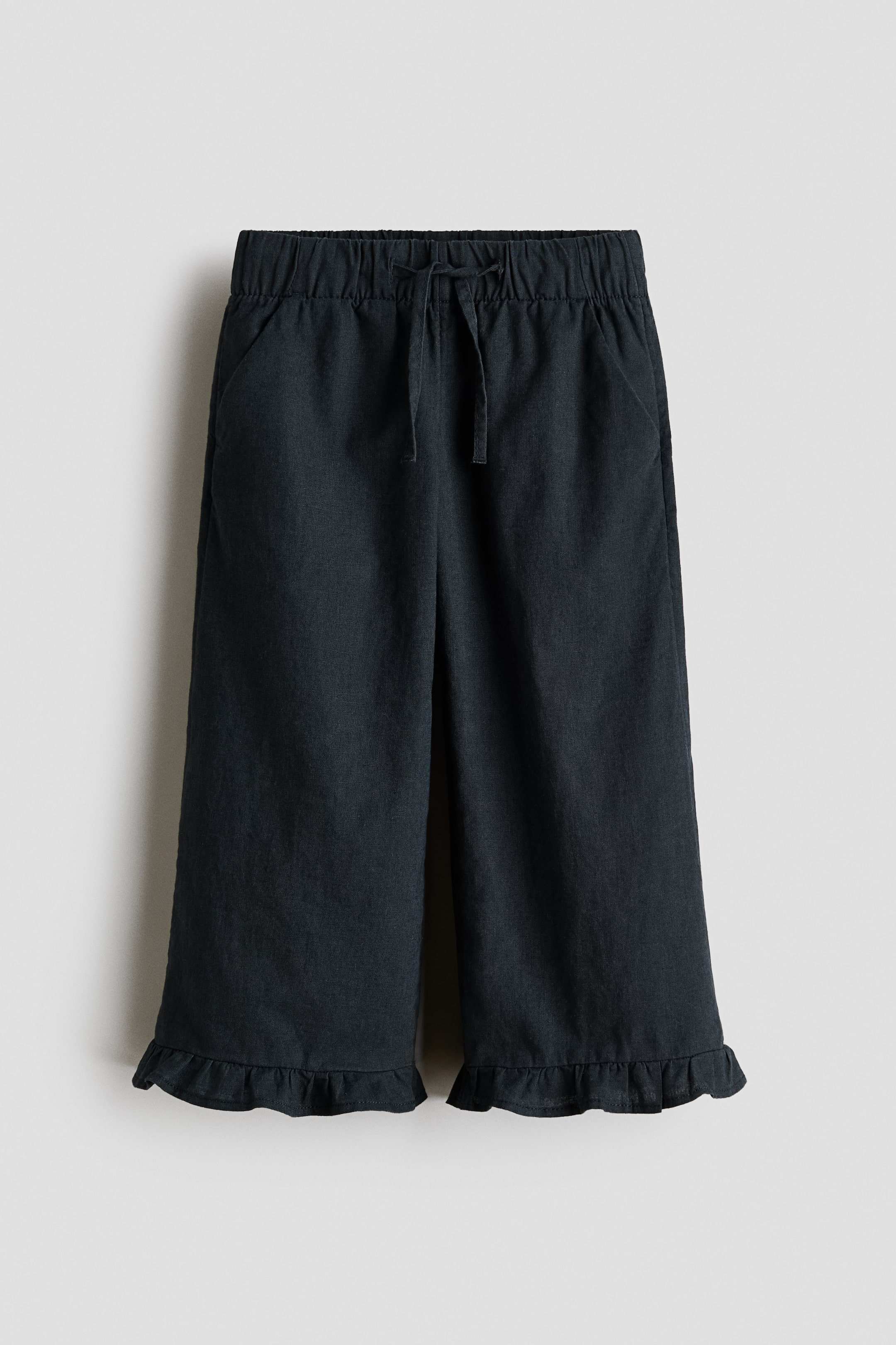 View larger image: Dark grey wide-leg cropped trousers are showcased from the front. They highlight an elasticated drawstring waistband with a tie, and two diagonal side pockets are positioned on the hips. The wide legs conclude with ruffled hems.
