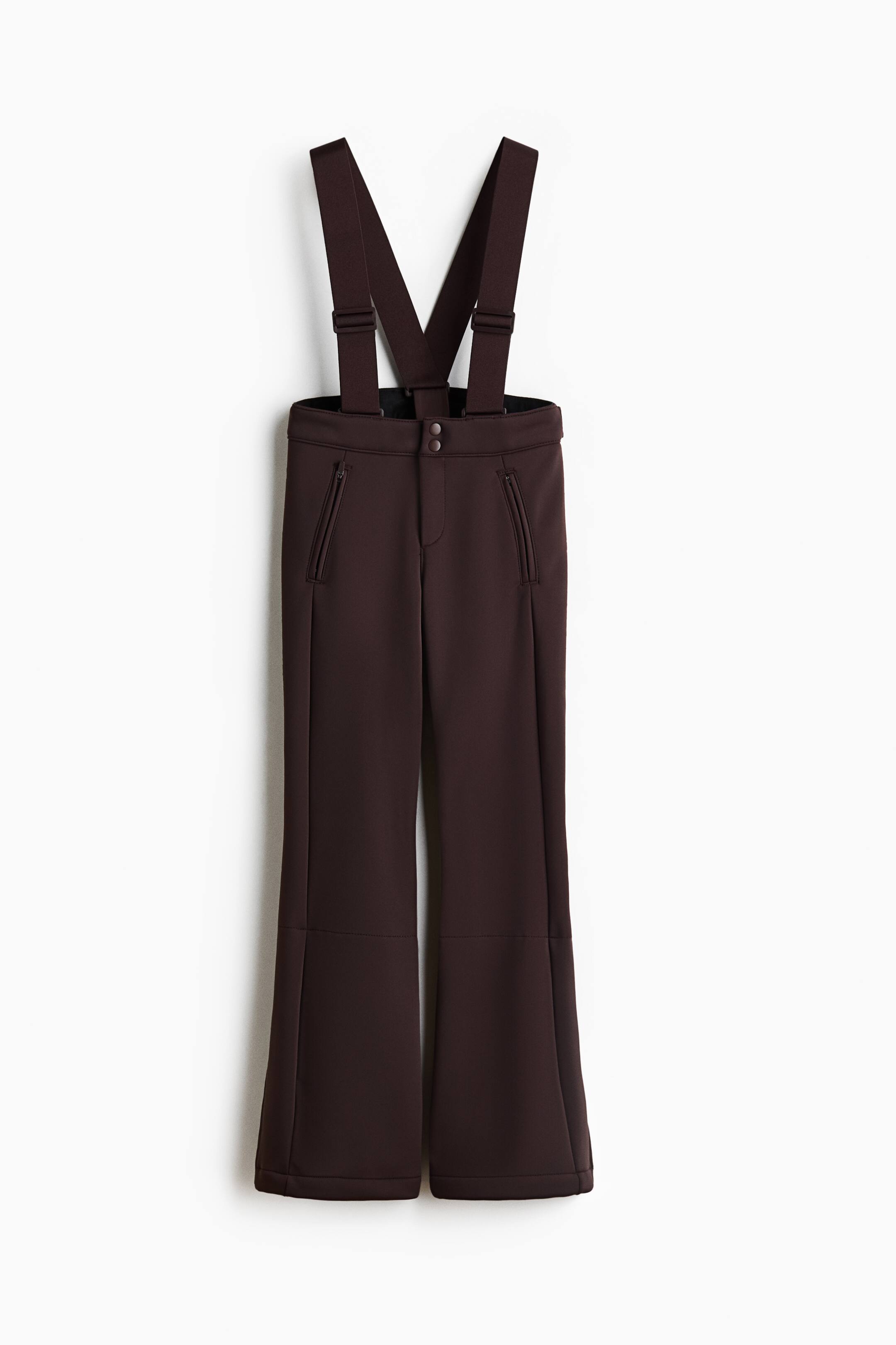 Ampliar la imagen: Estos pantalones de esquí color café oscuro están orientados hacia el frente y presentan tirantes anchos y ajustables sujetos a una pretina de tiro alto con cierre de doble botón a presión. Los pantalones muestran una silueta acampanada, con bolsillos frontales con cierre diagonal y costuras verticales que recorren cada pierna.