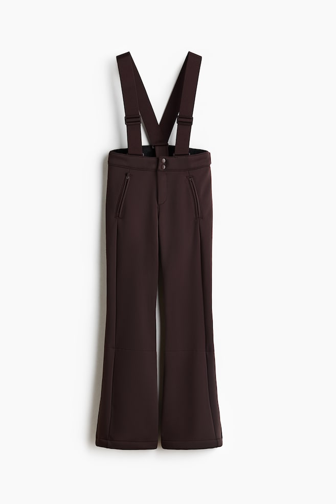 These dark brown ski trousers are facing front, featuring wide, adjustable suspenders attached to a high-rise waistband with a double snap-button closure. The trousers are showcasing a flared silhouette with diagonal zippered front pockets and vertical seams running down each leg. - 1