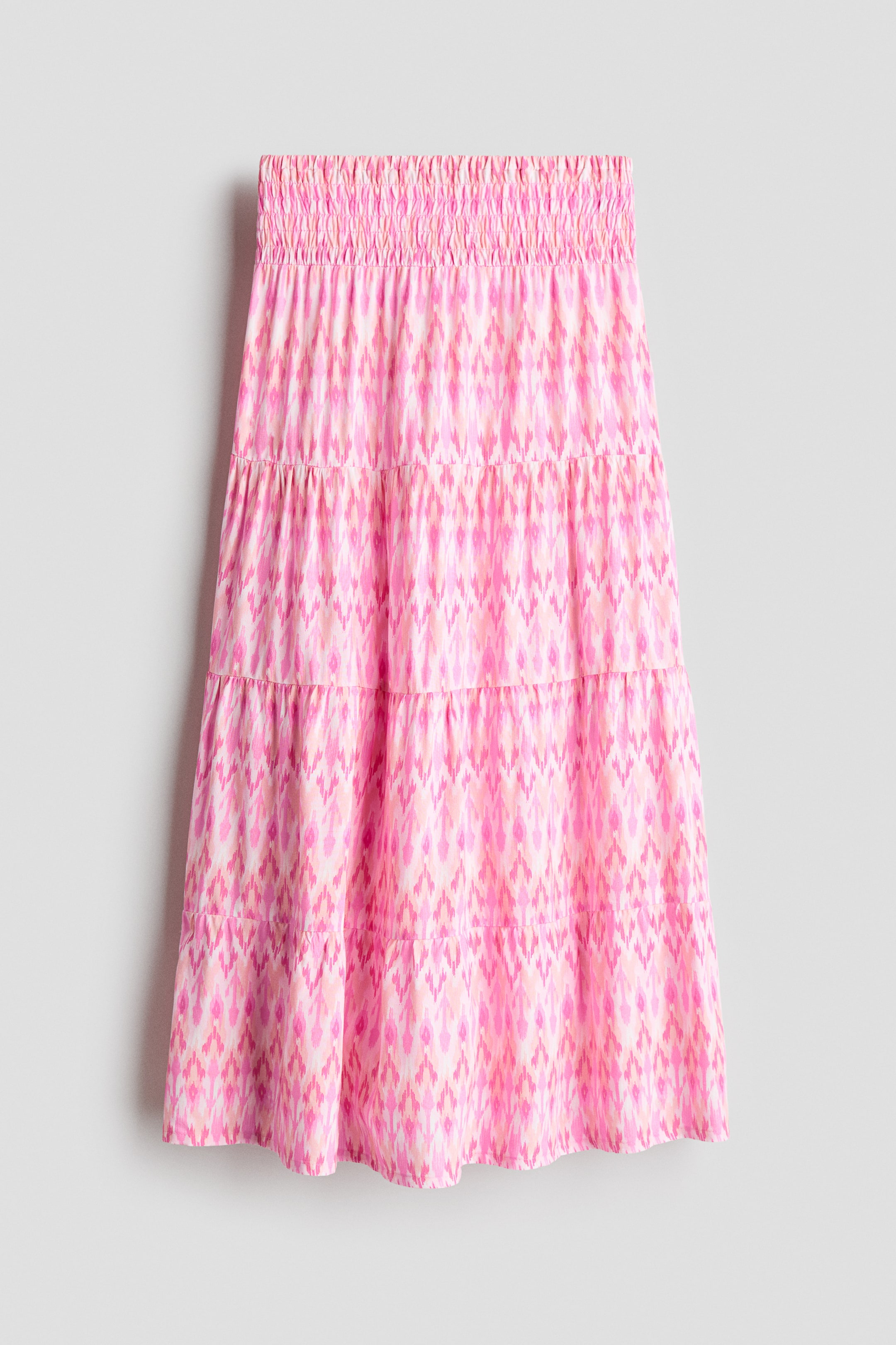 Größeres Bild anzeigen: Der leicht A-förmige, stufige Rock aus einem luftigen Gewebe ist vorne ausgerichtet und verfügt über einen gesmokten Bund. Er zeigt ein abstraktes geometrisches Muster in Rosa-, Pfirsich- und Weißtönen.