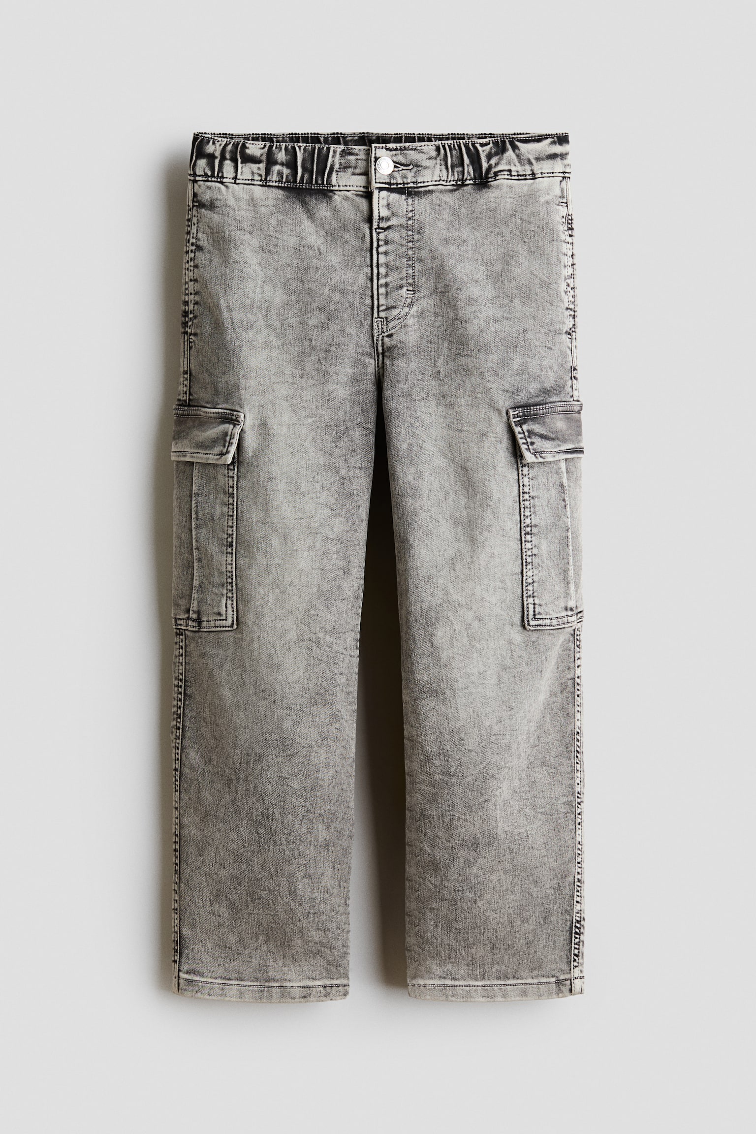 A pair of grey acid-wash denim cargo trousers is shown from the front, featuring a straight-leg silhouette and a gathered elasticated waistband with a single-button fastening. The trousers display flap cargo pockets on both sides of the thighs with prominent topstitching along the seams.