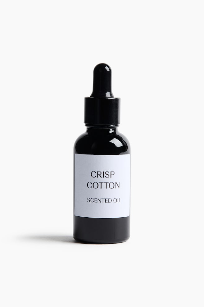 Um frasco conta-gotas de vidro preto estÃ¡ de pÃ© na vertical, virado para a frente, exibindo uma etiqueta retangular branca com o texto preto "CRISP COTTON" e "SCENTED OIL". O frasco apresenta uma tampa roscada preta integrada com um conta-gotas de bulbo de borracha. - 1