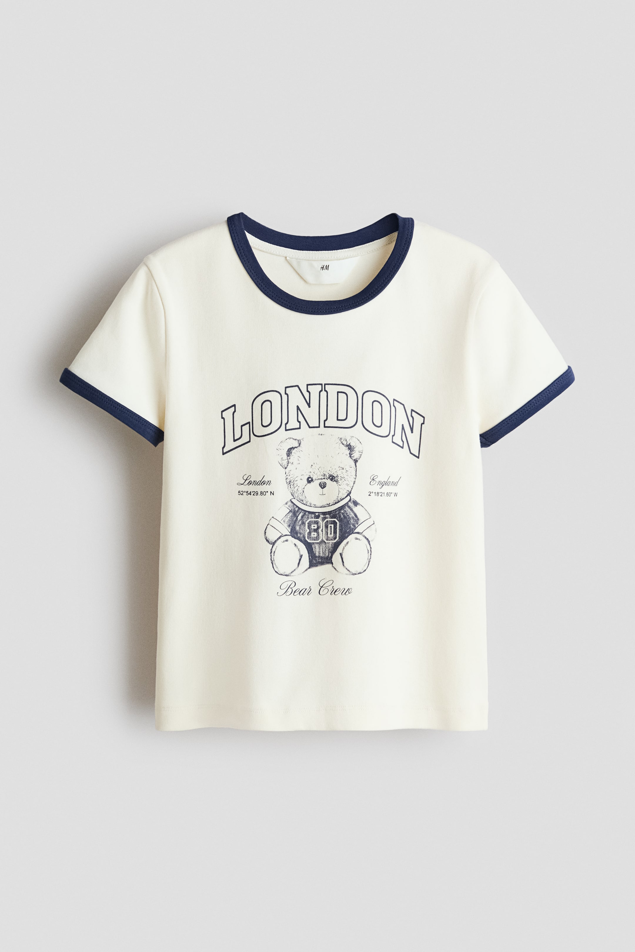 Zobacz większy obraz: Kremowy t-shirt posiada ciemnoniebieską lamówkę na okrągłym dekolcie oraz krótkich rękawach. Przód koszulki zdobi ciemnoniebieski nadruk graficzny z napisem "LONDON" w łuku nad misiem w koszulce z numerem "80", z napisami "London 52°54'29.80" N" oraz "England 2°18'21.60" W" po bokach misia, a poniżej znajduje się napis "Bear Crew".