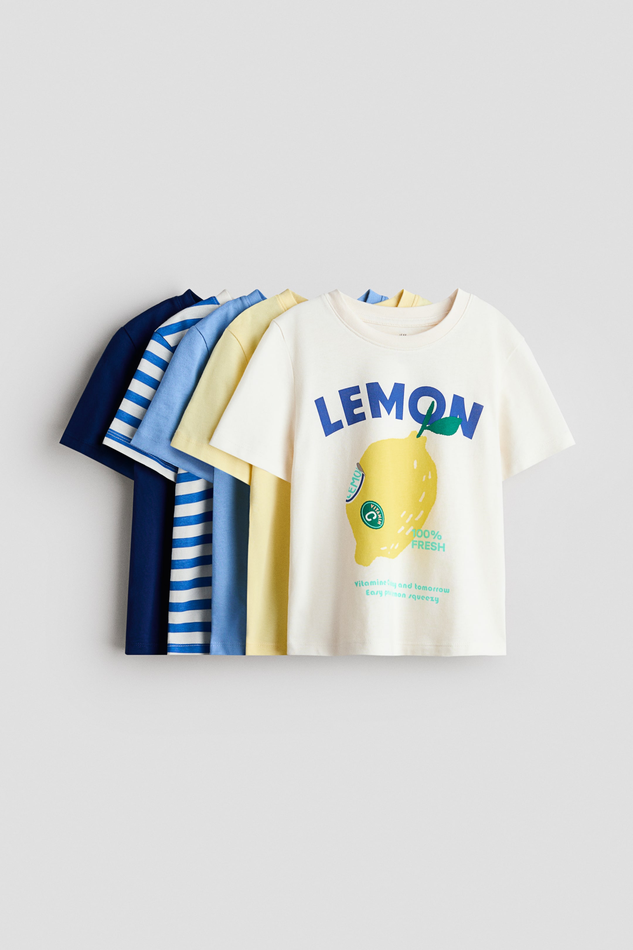 View larger image: A stack of five classic T-shirts crafted from soft cotton jersey features round, ribbed necklines and showcases a variety of colors and designs. The front T-shirt is displayed facing forward, exhibiting an off-white color with the word "LEMON" in blue and a yellow lemon graphic on the front. The other T-shirts come in solid light yellow, light blue, dark blue, and a dark blue and white stripe pattern positioned behind it.