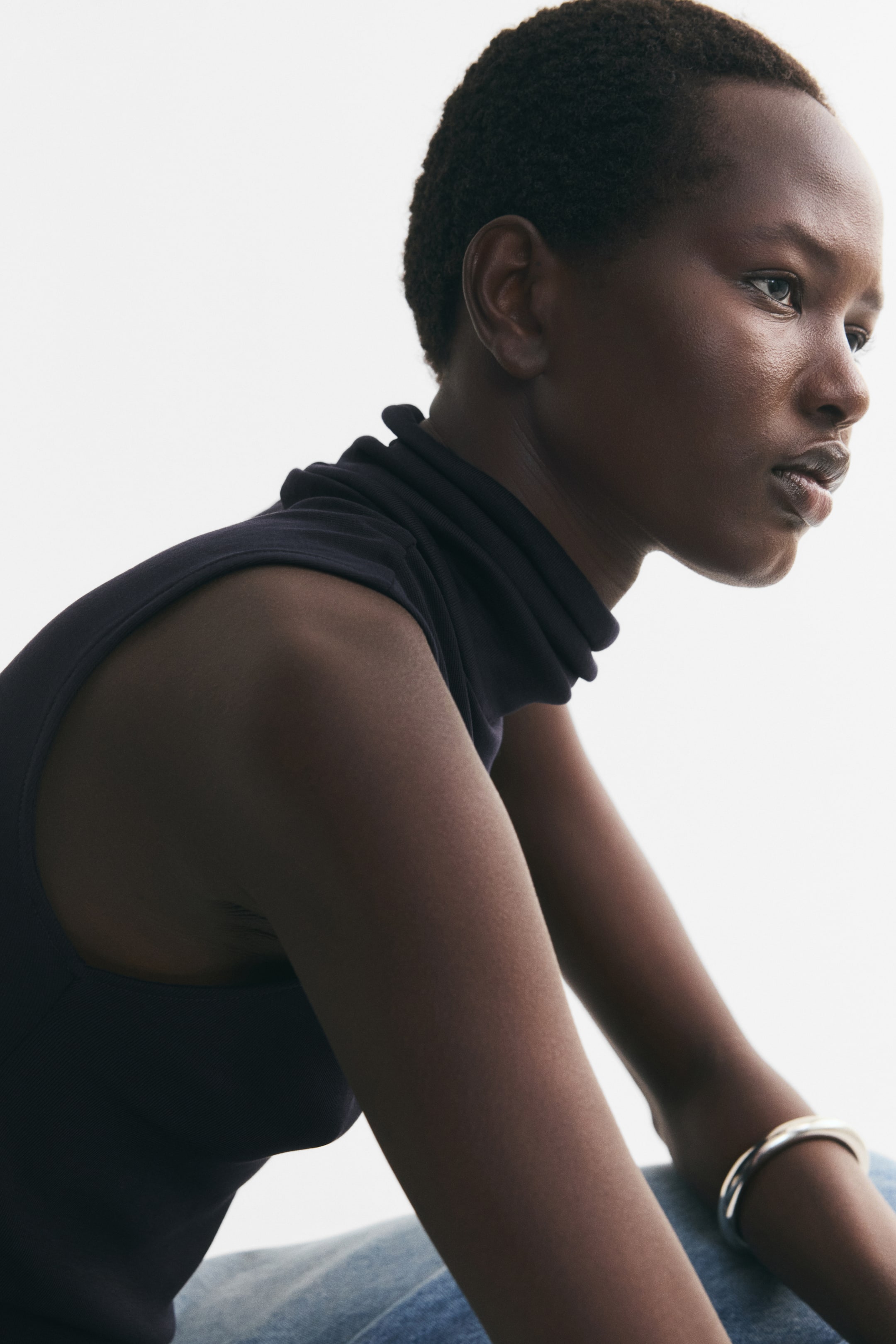 View larger image: A sleeveless, dark navy turtleneck top in a fine knit is being worn by the model, shown in profile. The top is styled with light-wash denim jeans and a silver-toned cuff bracelet.