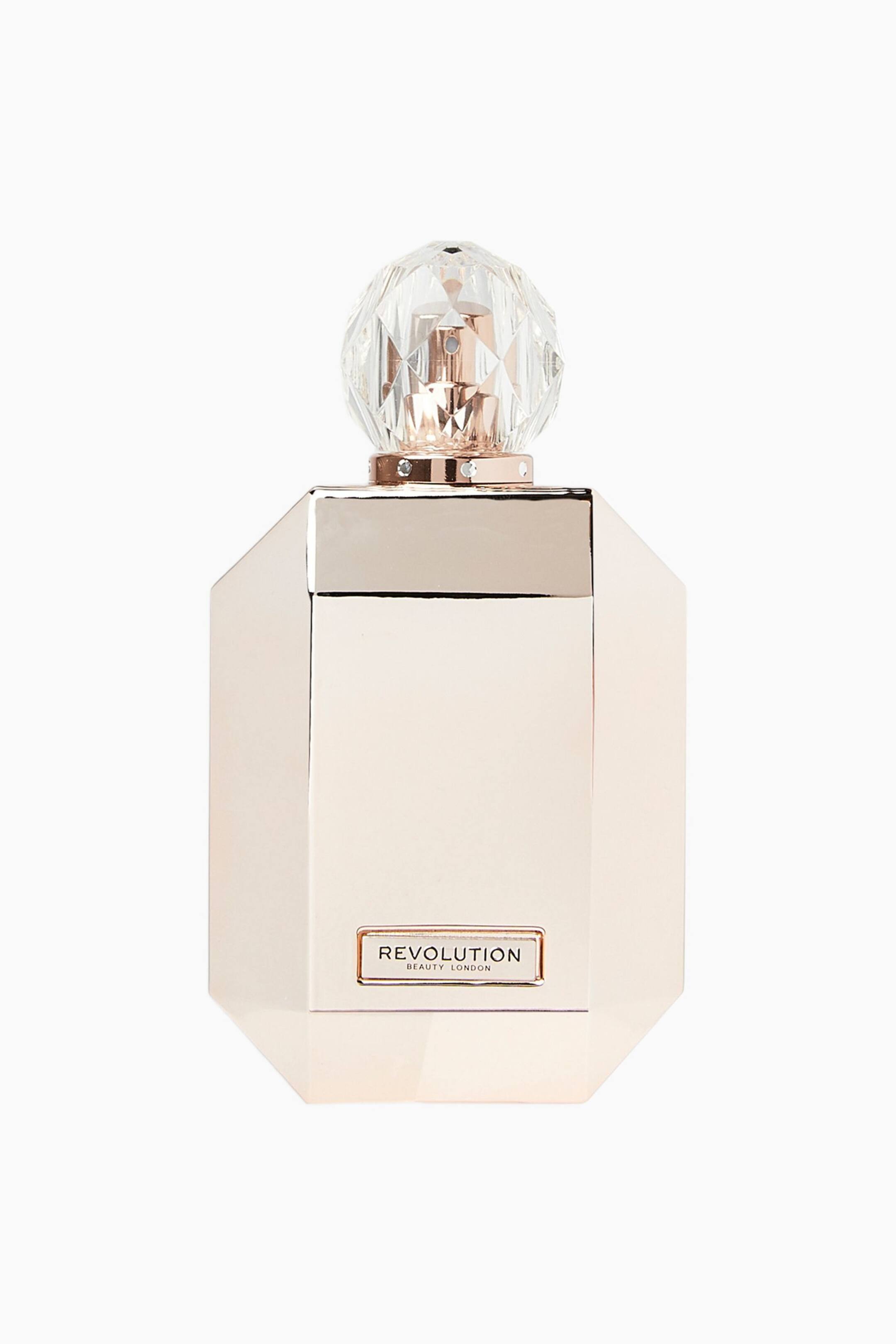 Größeres Bild ansehen: Diese Parfümflasche steht aufrecht und zeigt eine frontale Ansicht ihres achteckigen, metallisch roségoldenen Körpers mit einer kleinen rechteckigen Plakette nahe der Basis, auf der „REVOLUTION BEAUTY LONDON“ steht. Sie wird von einer klaren, facettierten, kristallähnlichen Kugelkappe gekrönt, die auf einem metallischen Kragen mit kleinen Nieten ruht.