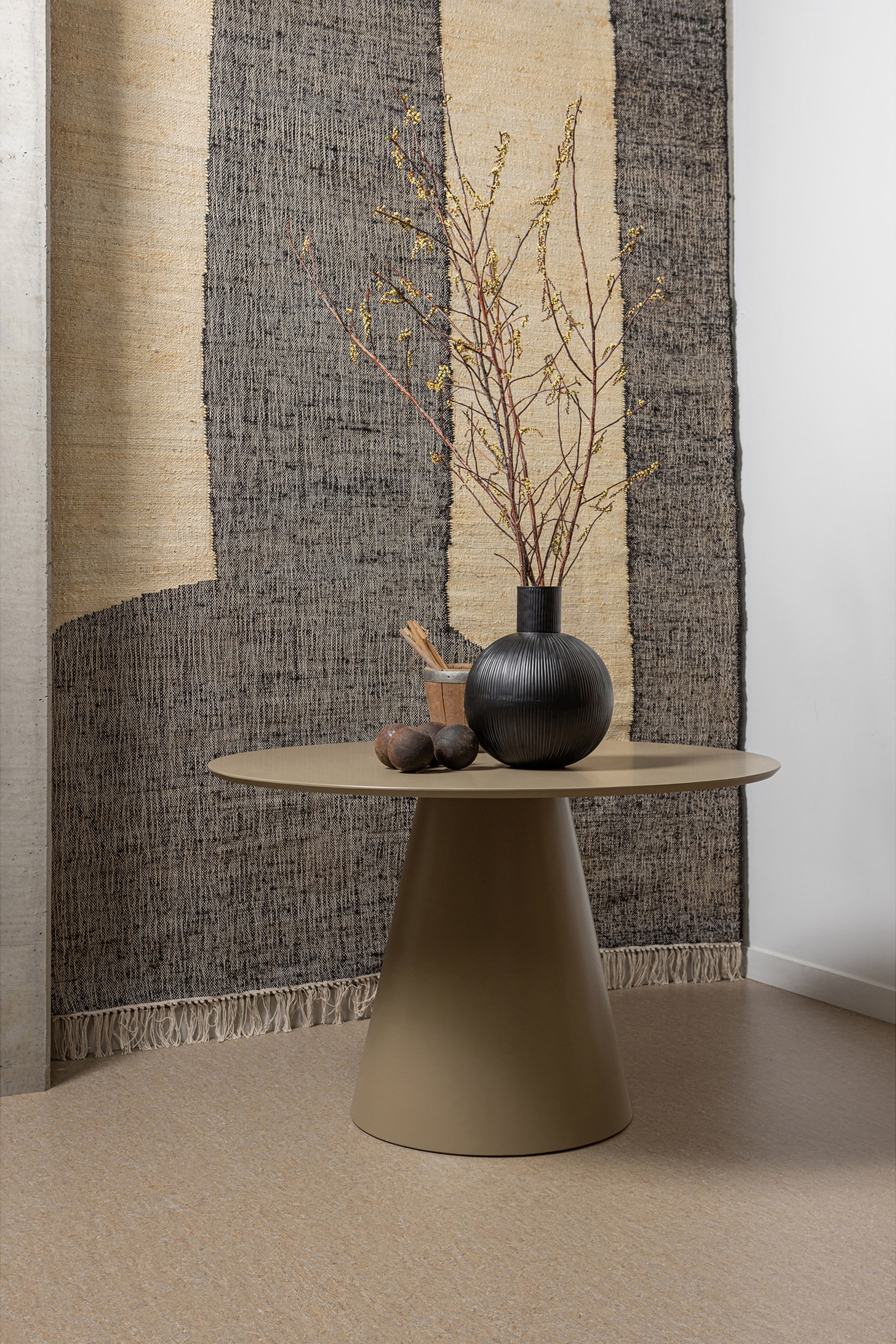 Grotere afbeelding bekijken: Een gedempte beige tafel toont een ronde bovenkant en een brede, taps toelopende basis, met een zwarte geribbelde vaas waarin takken en andere decoratieve items op het oppervlak rusten.