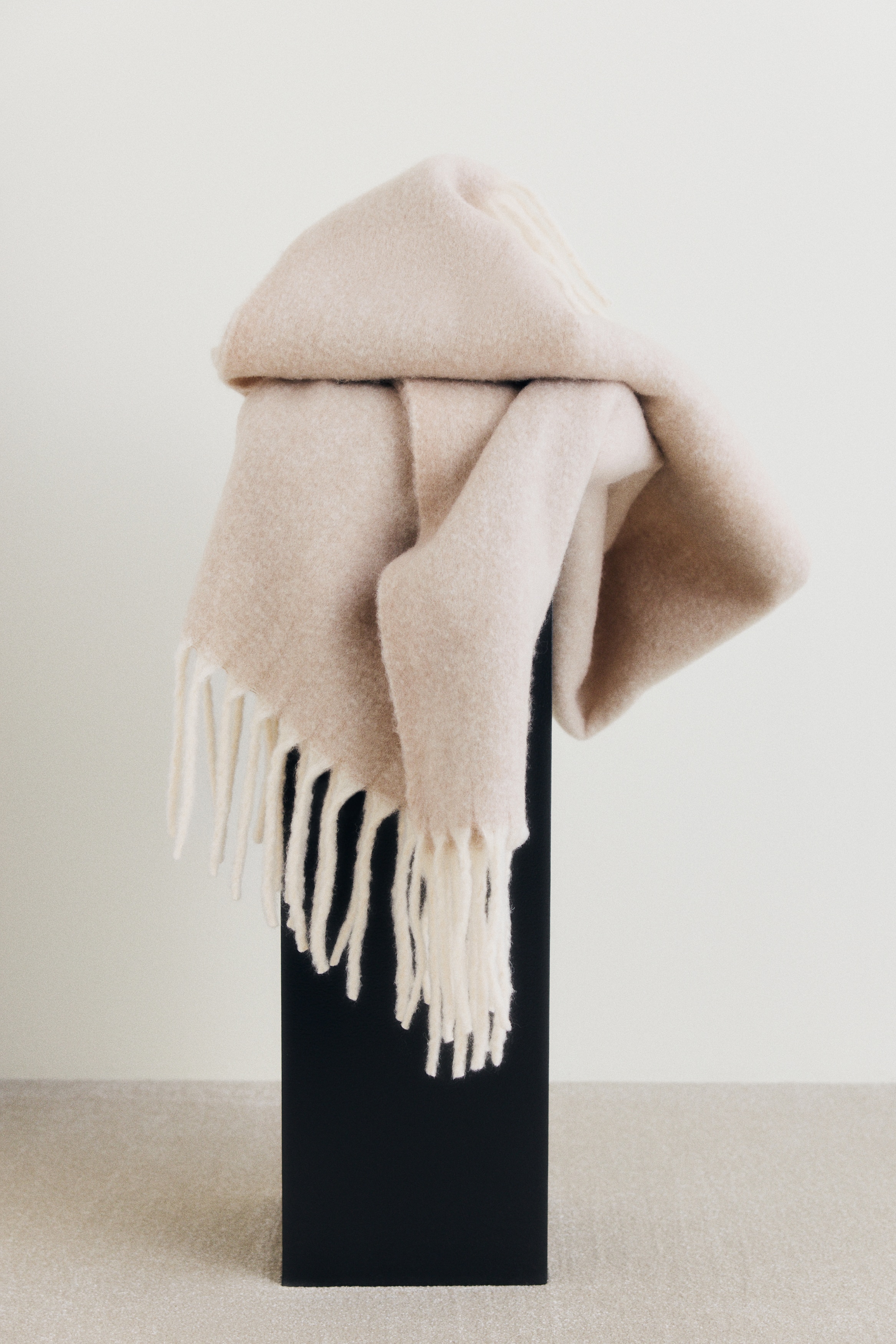 A light beige blanket is folded neatly into a rectangular shape, showcasing a soft, brushed texture across its surface. A contrasting cream-coloured fringe is visible along the bottom edge of the folded blanket.