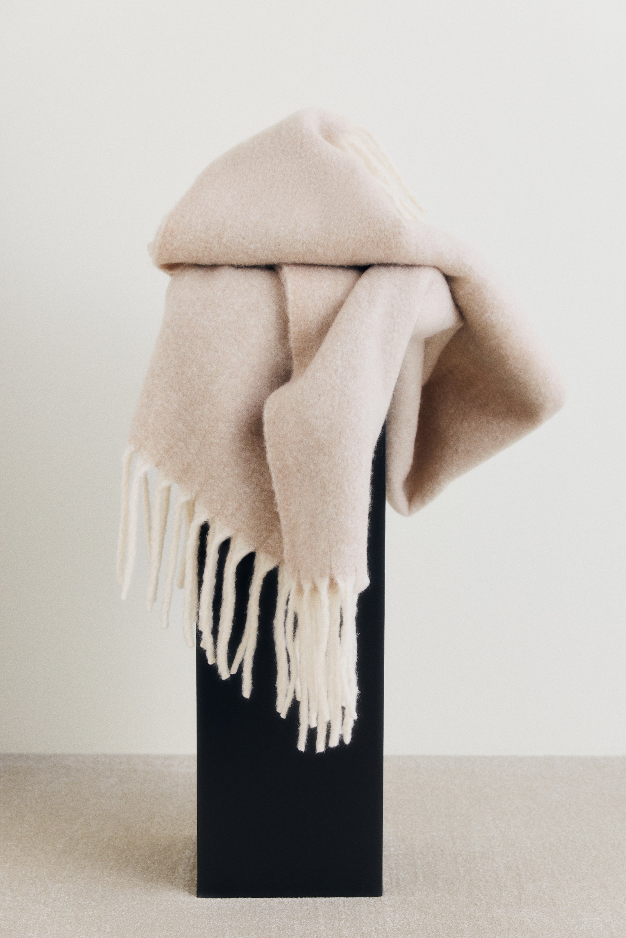 View larger image: A light beige wool-blend blanket with a soft brushed finish is draped over a black pedestal, showcasing its thick off-white fringe hanging along the bottom.