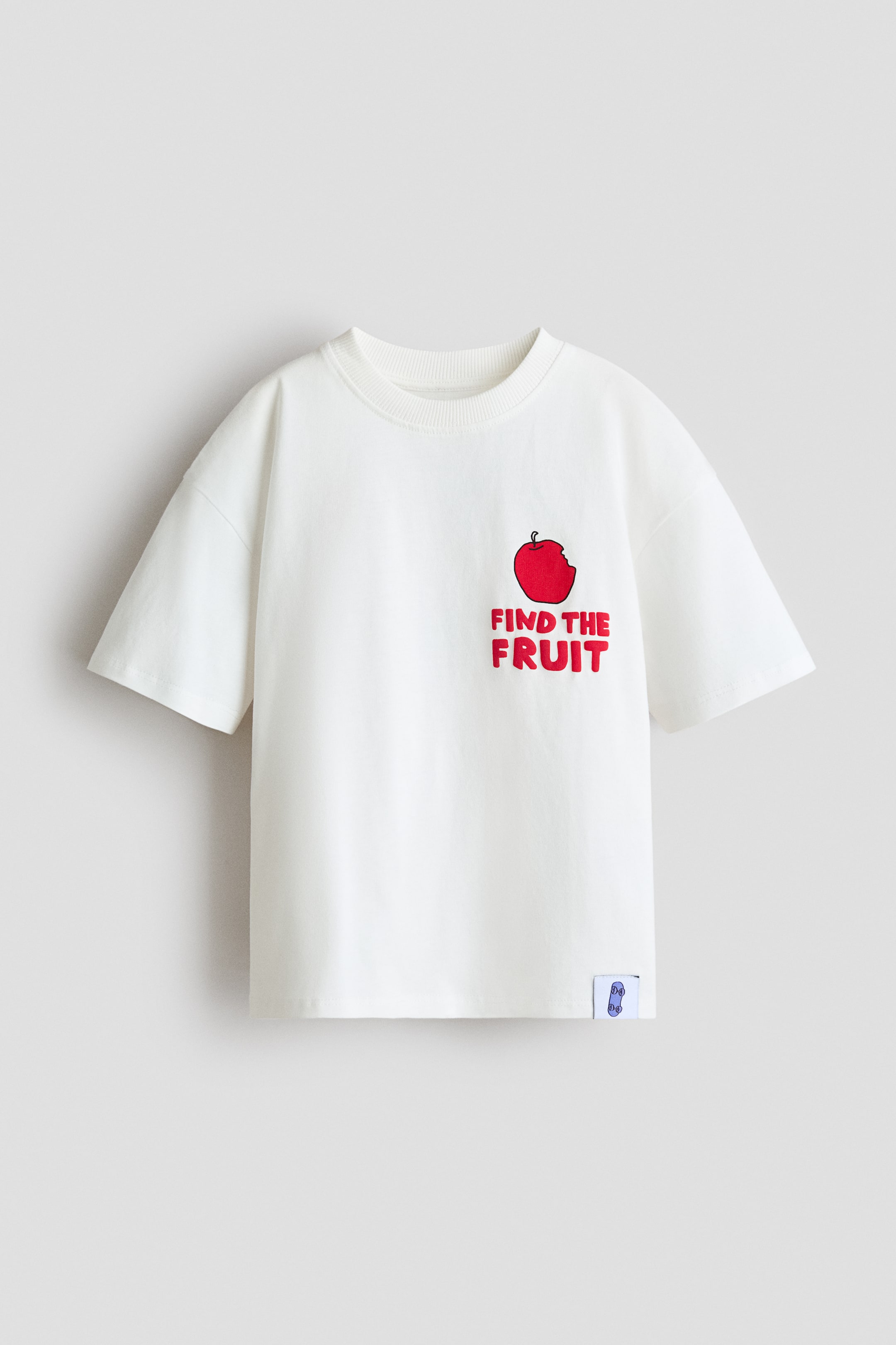View larger image: A loose-fit t-shirt in off-white cotton jersey is showcasing its front view, featuring a round, rib-trimmed neckline and dropped shoulders. It displays a red apple graphic with a bite taken out and the text "FIND THE FRUIT" on the front left chest, along with a small, purple and white graphic label visible on the bottom left hem.