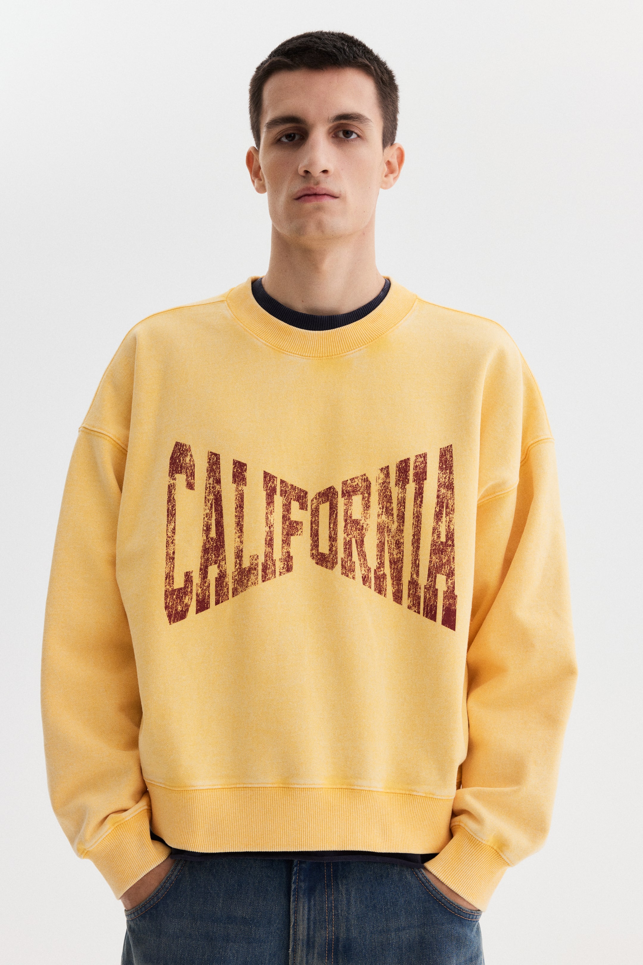 View larger image: A relaxed-fit yellow sweatshirt features a distressed dark red "CALIFORNIA" graphic across the front in an arched shape. It has a ribbed crew neck, ribbed cuffs, and a ribbed hem, and is worn over a dark blue T-shirt and partially over blue jeans.