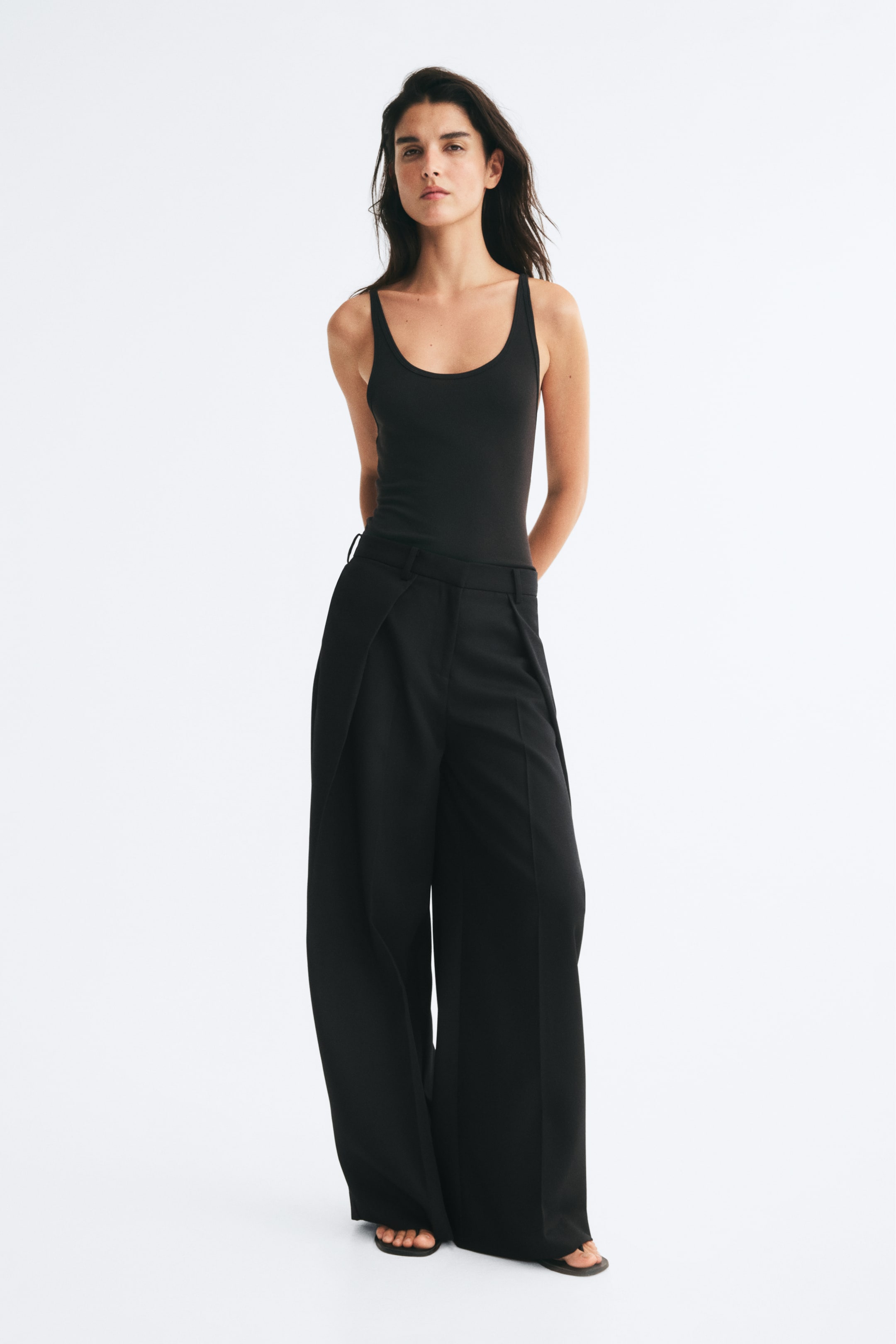 Grotere afbeelding bekijken: Zwarte loose-fit broek met wijde pijpen, wikkelvoorzijde en persplooien aan de voorkant, gedragen door het model. De broek is gestyled met een aansluitende zwarte tanktop.