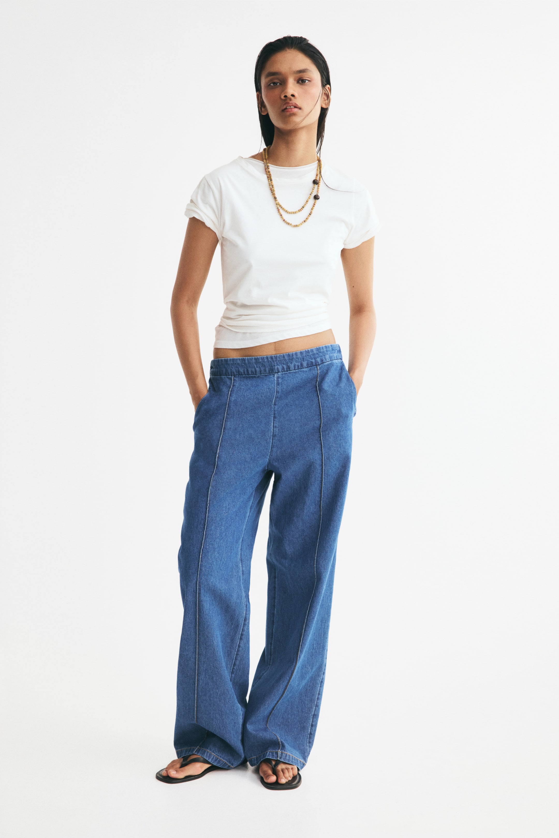 View larger image: The model is wearing relaxed-fit blue denim trousers. The trousers feature a flat elasticated waistband, discreet side pockets where the model’s hands are resting, and straight legs with sewn-in creases down the front.