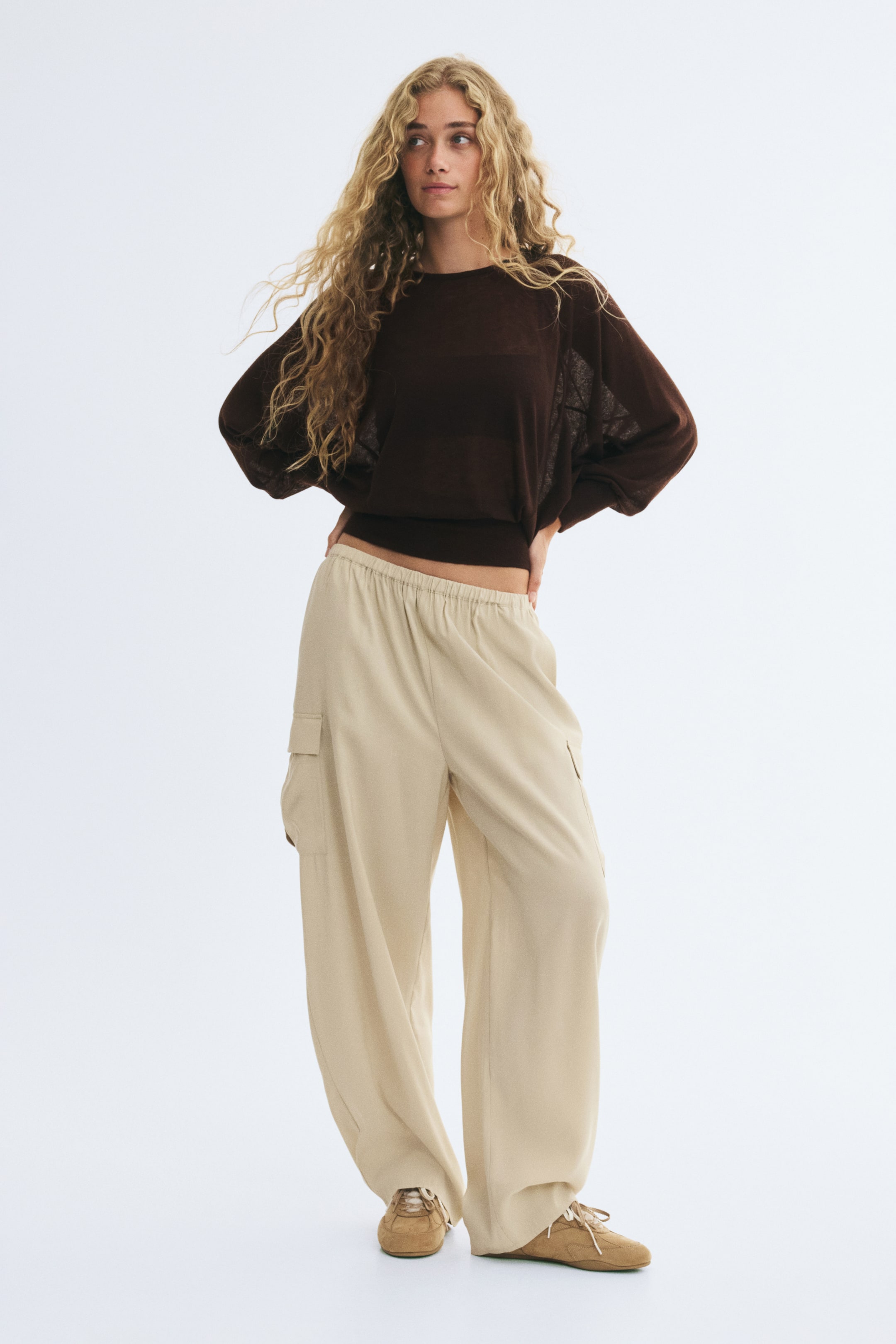 Se større billede: En mørkebrun, let strikket crewneck-sweater med løstsiddende ærmer bæres af modellen og er stylet med beige cargo-bukser med brede ben, elastisk talje og praktiske sidelommer. Modellen står med hænderne på hofterne og fremhæver outfitets løse fald og afslappede pasform.