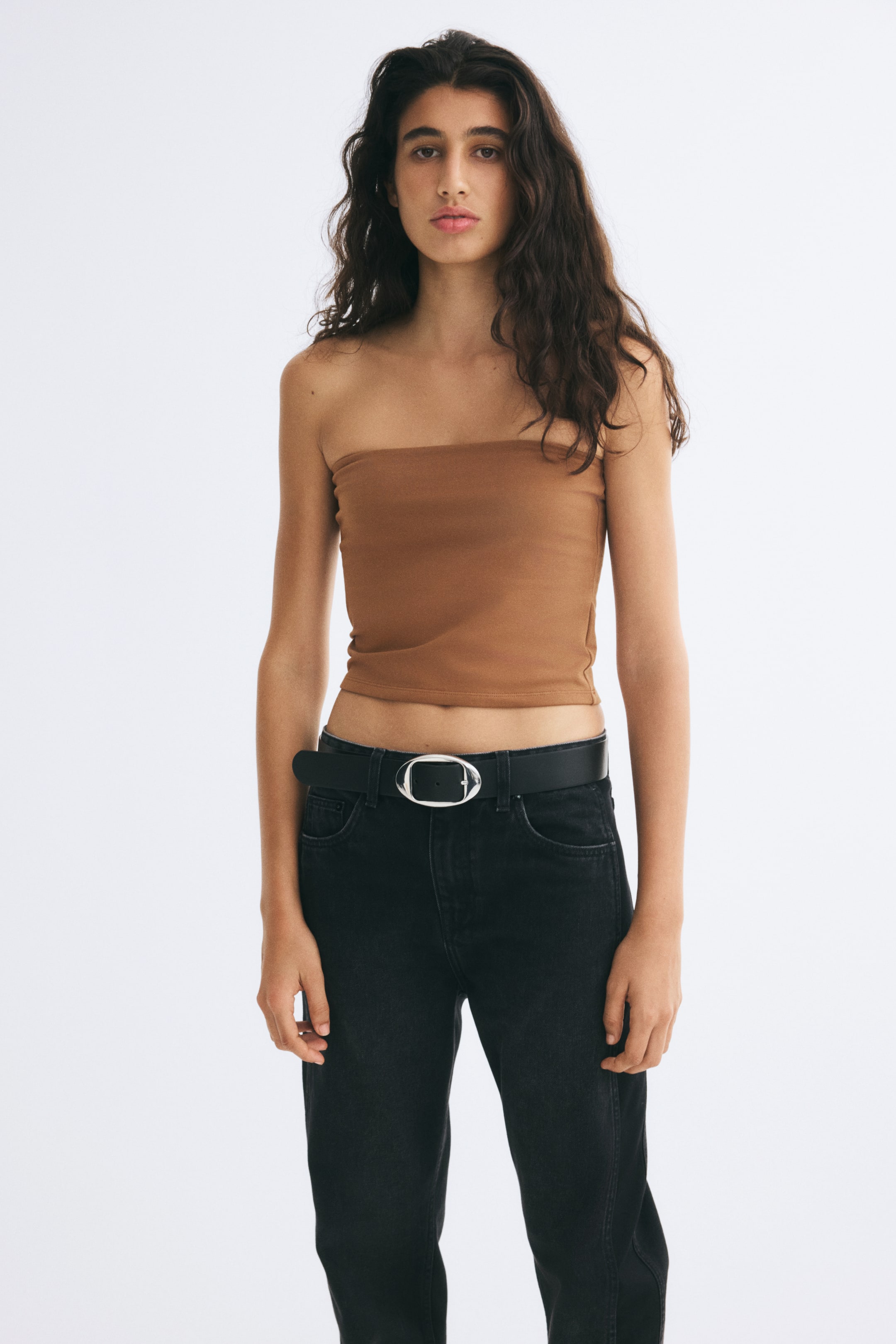 View larger image: The model is wearing a brown, fitted bandeau top with a strapless, straight-cut design. The top is styled with black jeans and a black belt with a silver buckle.