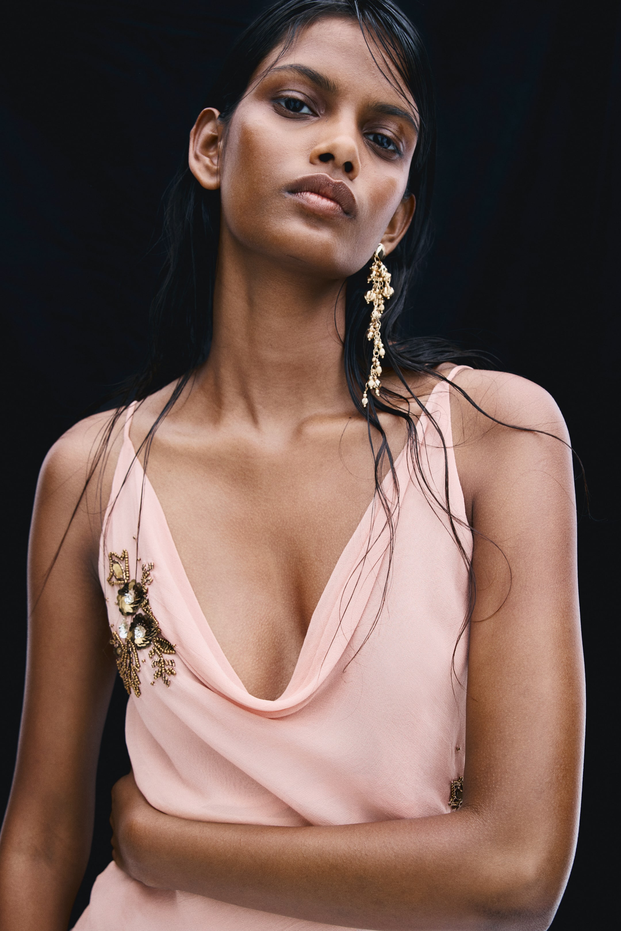 View larger image: The model is wearing a light pink top featuring a draped cowl neckline, thin spaghetti shoulder straps, and a beaded floral embellishment on the front. She is posing with one arm crossed over her waist, showcasing the top’s lightweight, fluid fabric.