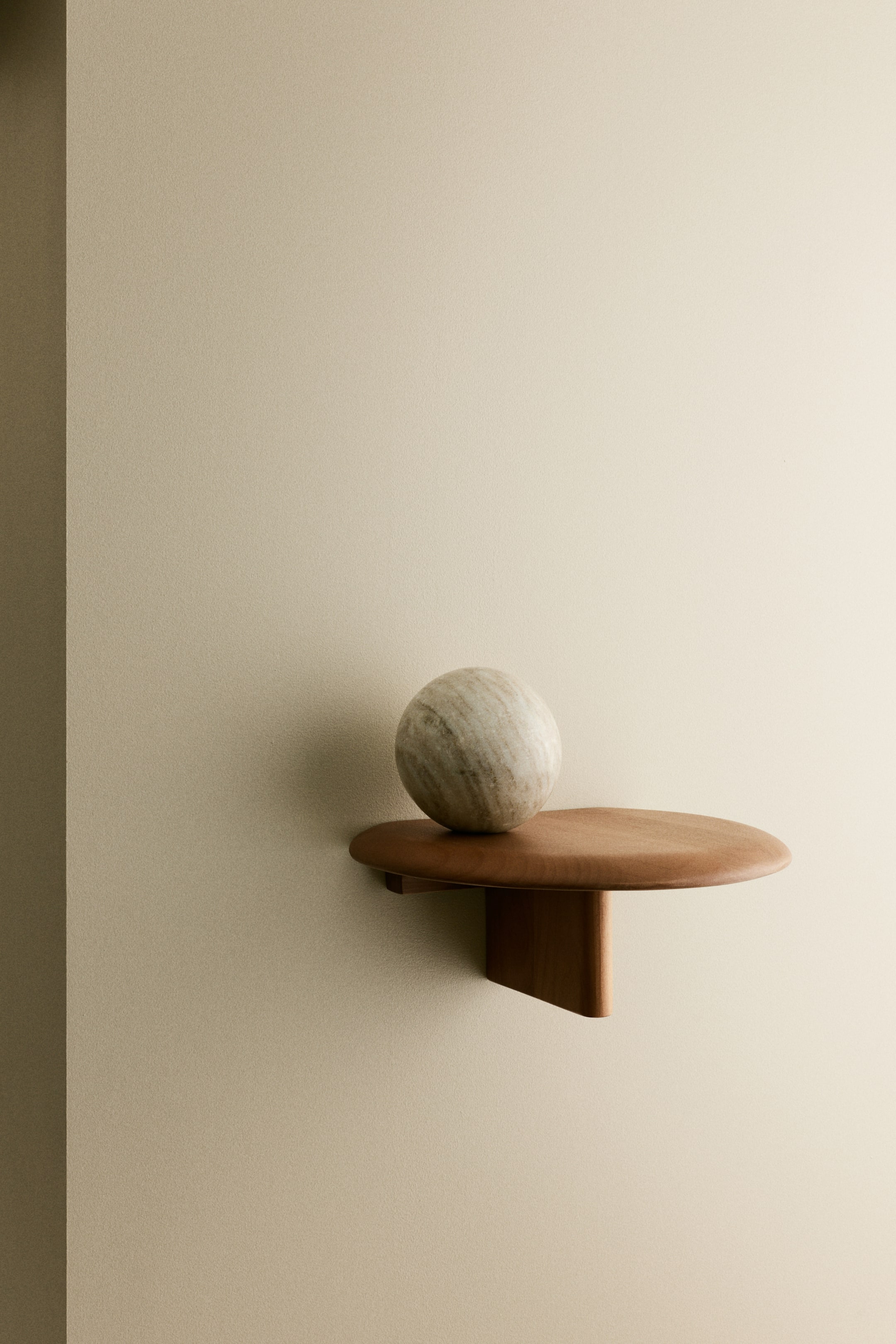 Grotere afbeelding bekijken: Een aan de muur gemonteerde, ronde houten plank steekt uit de muur en heeft een rijke bruine afwerking met één ondersteuningsstuk. Een bolvormig marmeren object ligt gecentreerd op de plank en vertoont subtiele witte en grijze aders.