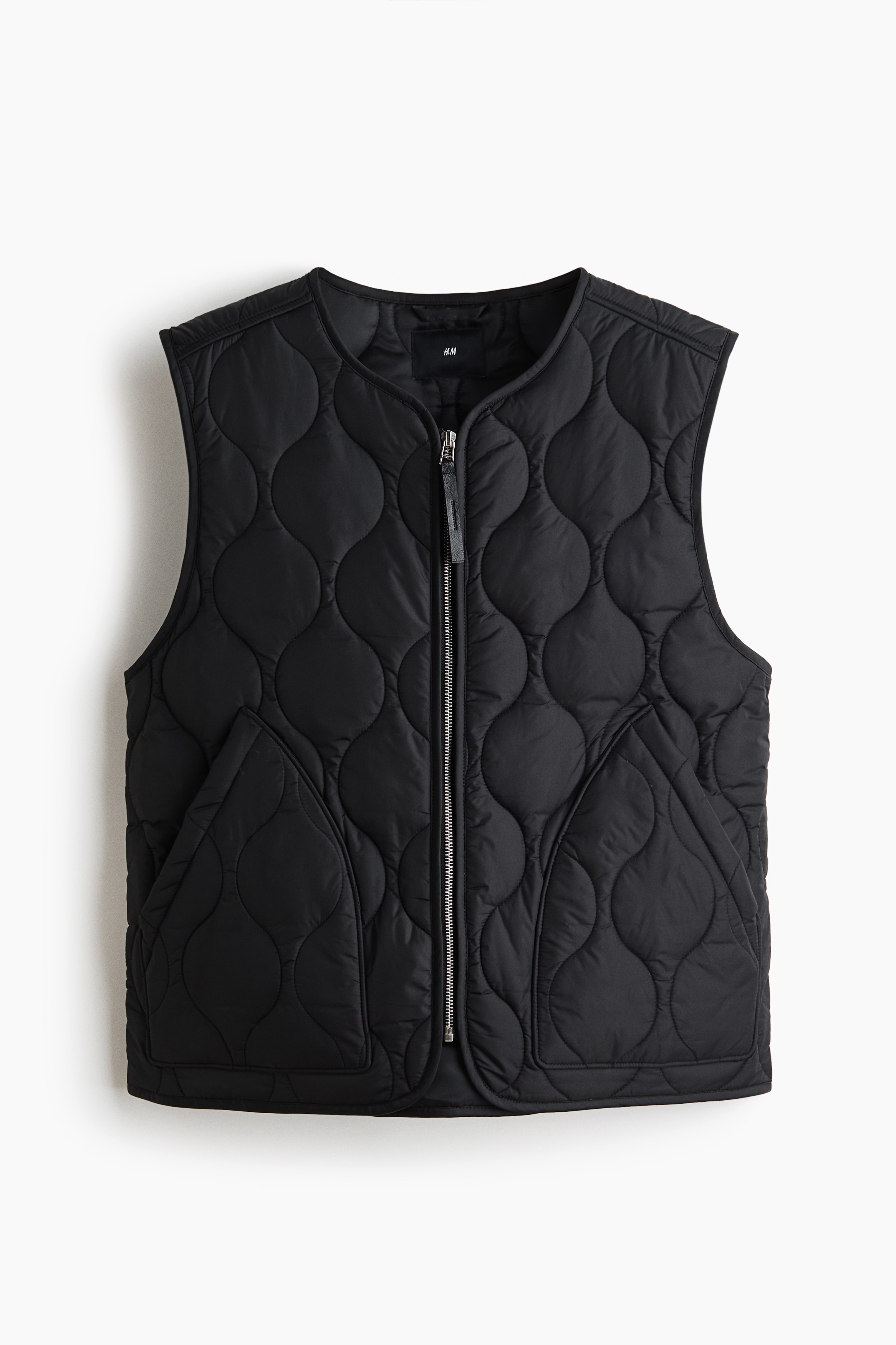 A black quilted nylon vest with a round neckline and front zipper is being worn by the model over a white t-shirt. The model is holding their hands inside the vest's front patch pockets.