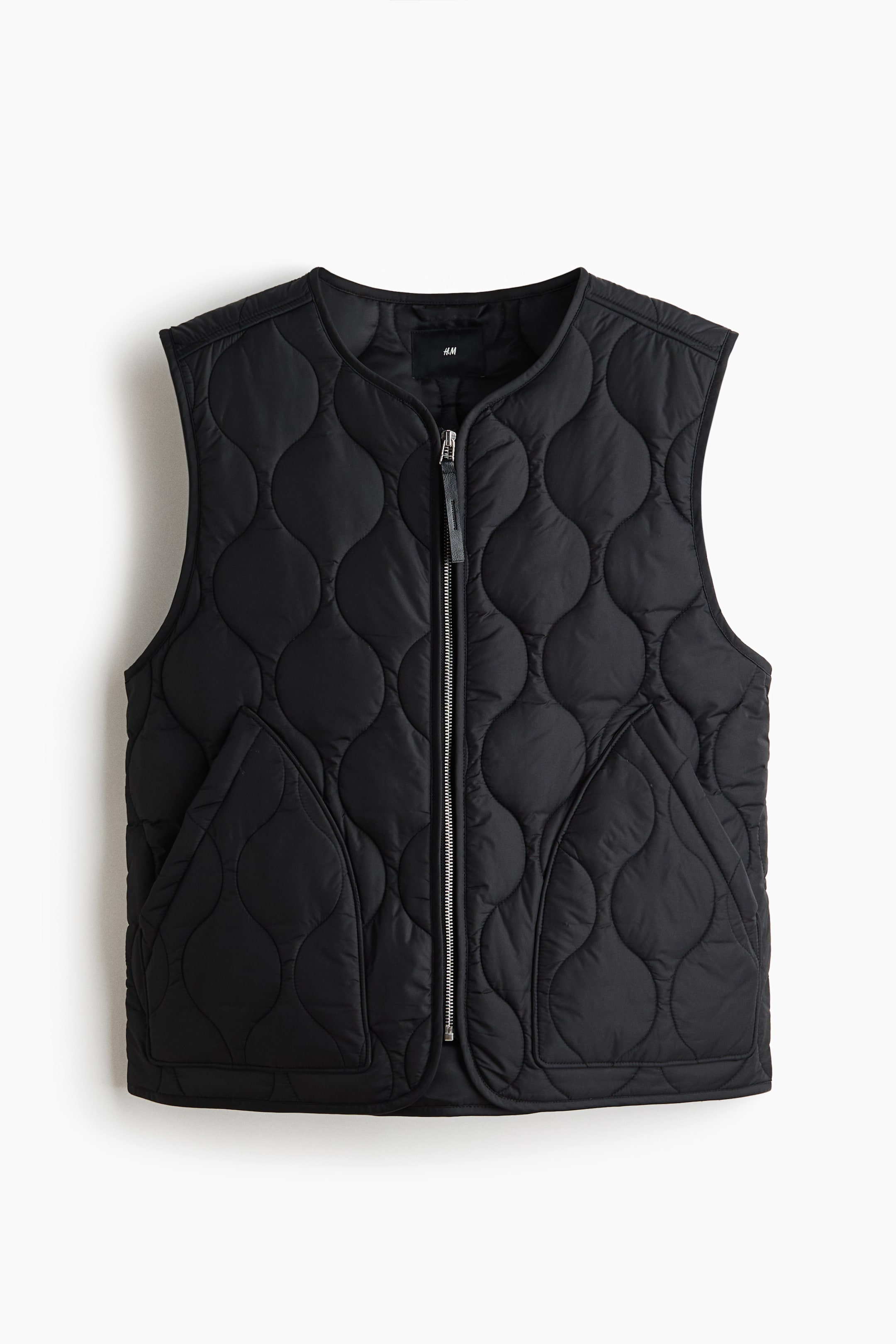 View larger image: A black quilted vest is shown front-facing, featuring a scalloped quilting pattern across its surface. A silver-toned zipper runs down the center front with a black pull tab, and two angled slash pockets are positioned on the lower front.