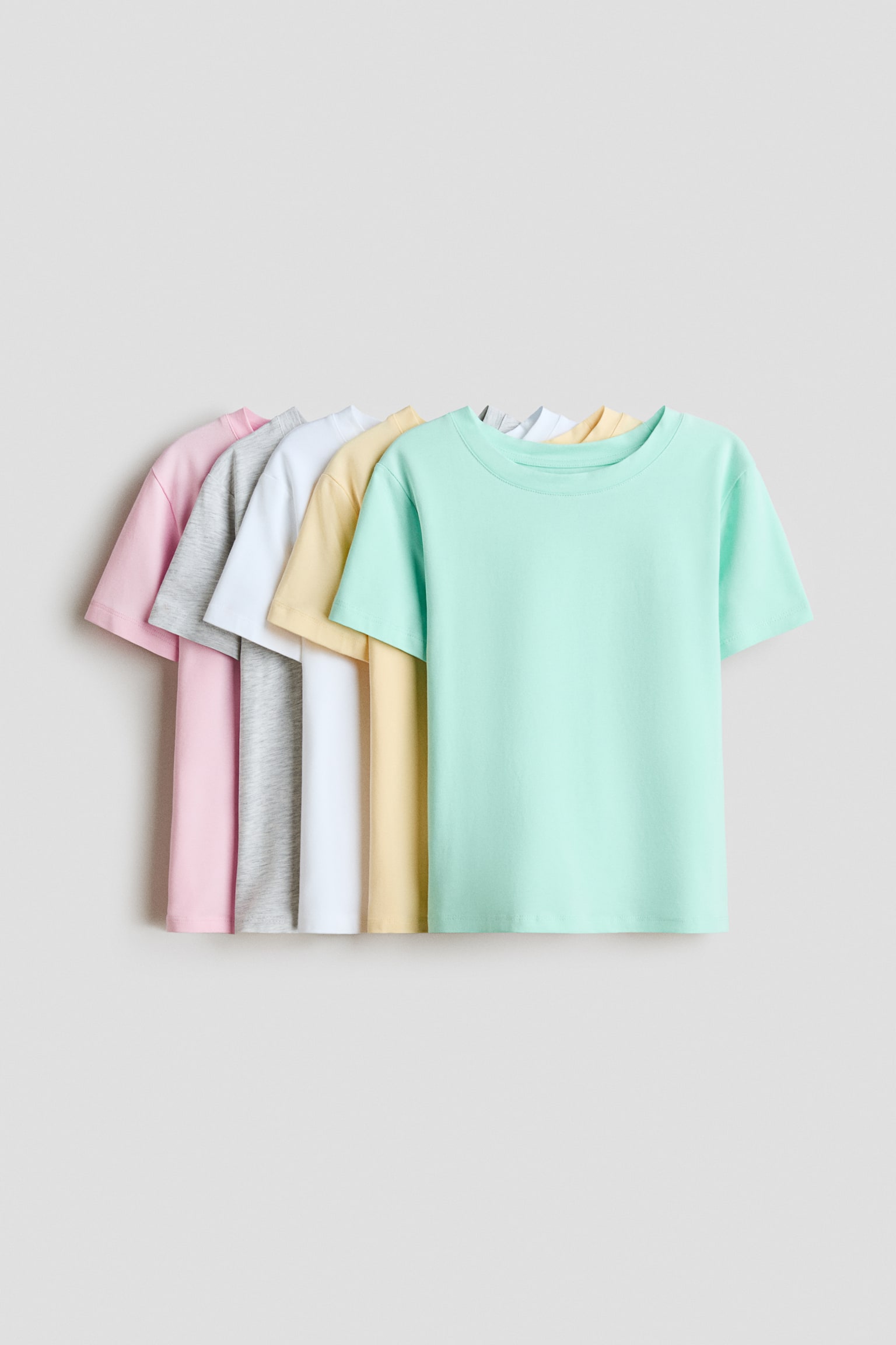 Five cotton jersey t-shirts are overlapping in a row facing front, showcasing a mint green t-shirt in the foreground followed by pastel yellow, white, heather grey, and light pink t-shirts. Each t-shirt features a round crew neckline with a narrow trim and short sleeves.