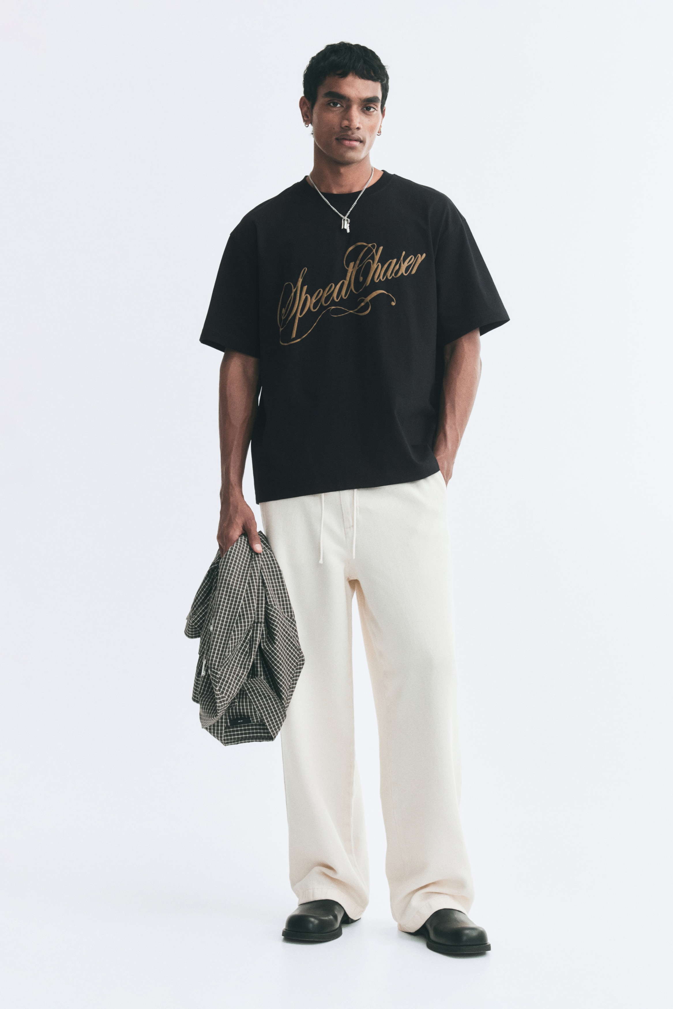 A black cotton jersey T-shirt is shown from the front, featuring a relaxed fit, a ribbed crew neckline, dropped shoulders, and a straight hem. The front displays a metallic gold cursive graphic reading "Speed Chaser" across the chest.