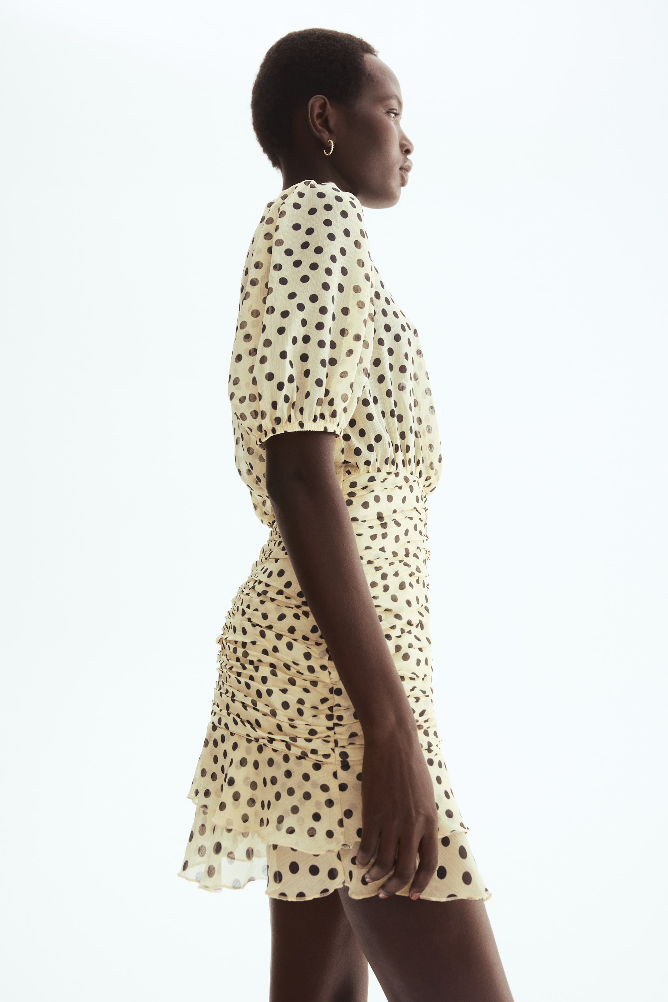 View larger image: A light yellow polka dot dress is displayed from the side, showcasing puffed short sleeves with elasticated cuffs, a ruched waist, and a ruffled hem.