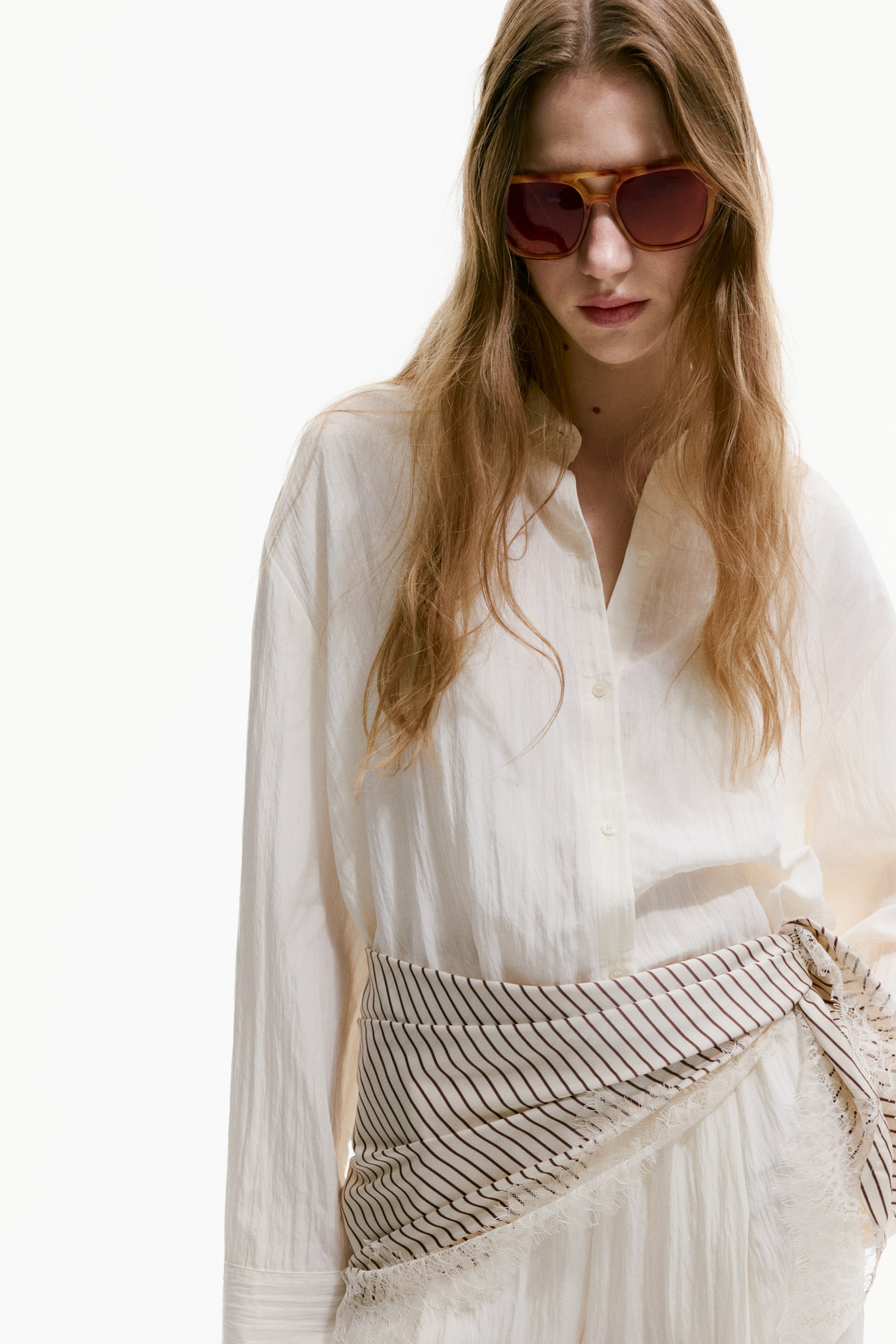 הצגת תמונה גדולה יותר: חולצה לבנה עם שרוולים ארוכים ובגזרה רפויה, בעלת מרקם מקומט, נלבשת עם פתיחה חלקית של הכפתורים ושרוולים מכופתרים. צעיף מפוספס עם קצוות פרנזים נכרך באלכסון סביב המותן, מונח מעל החולצה וחצאית לבנה עם מרקם.