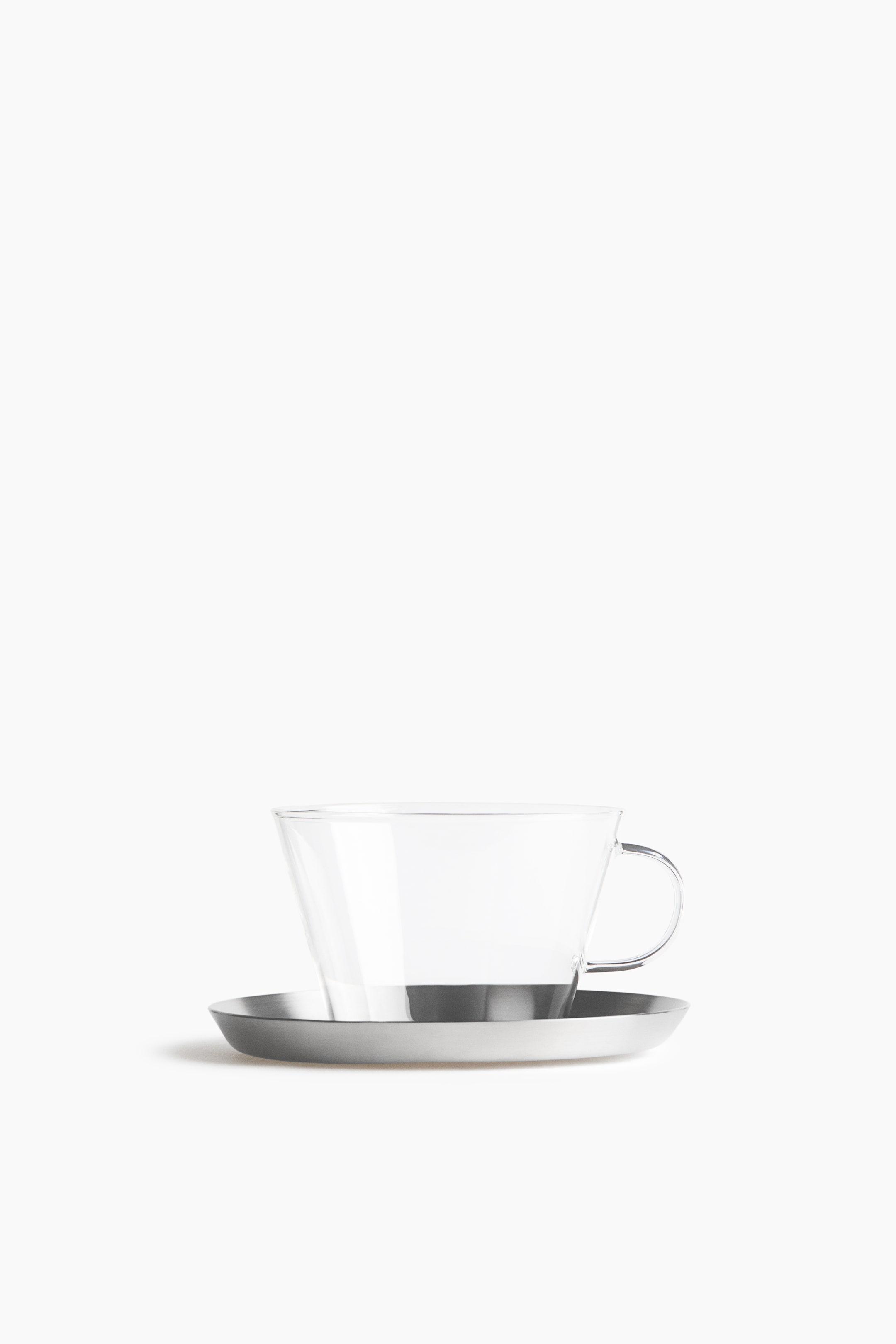 View larger image: A clear glass cup with a flared rim and a curved side handle is resting on a round brushed-metal saucer, shown from a front-facing angle.