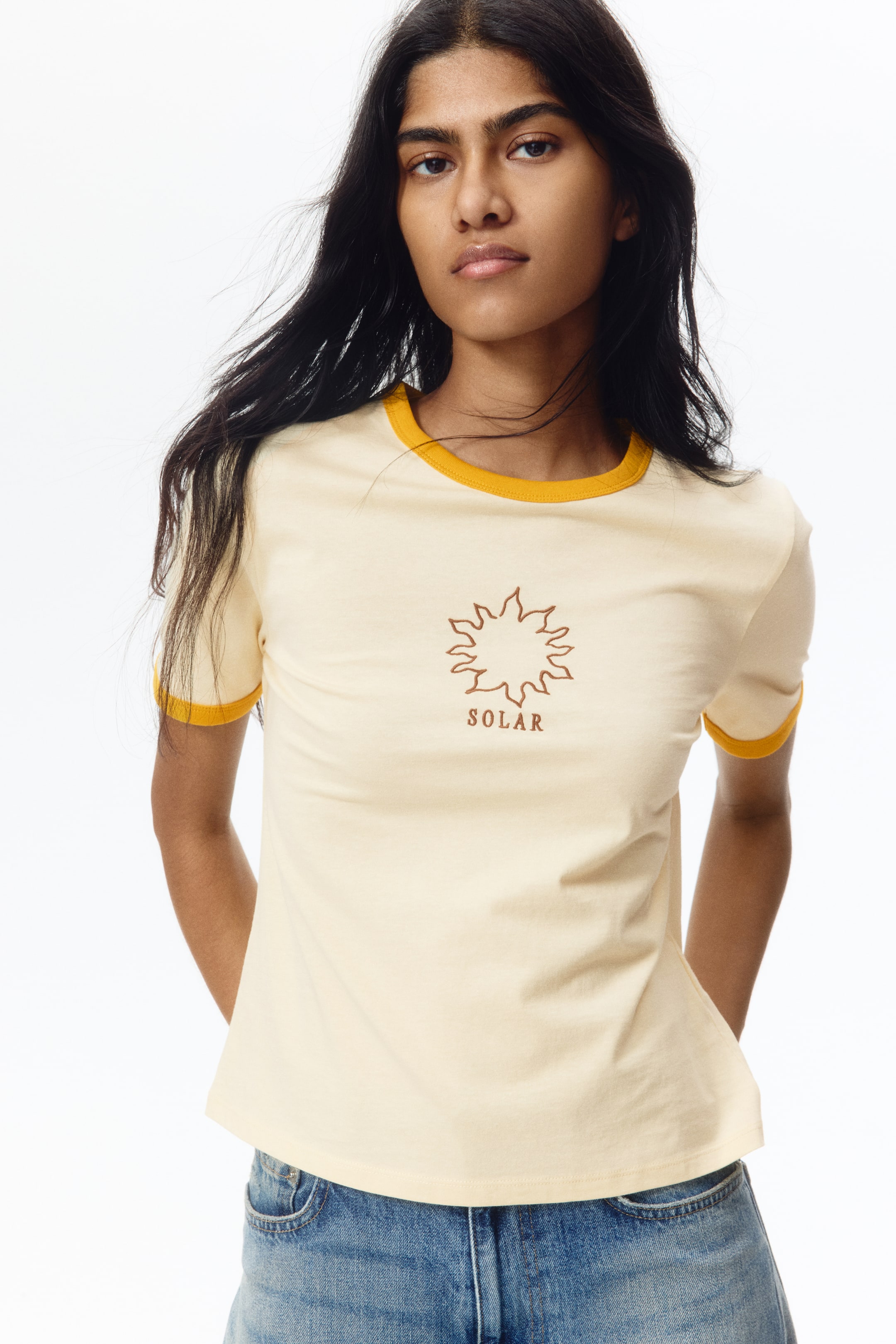 View larger image: A pale yellow ringer t-shirt is being showcased, featuring a round neckline and short sleeve cuffs with contrasting mustard yellow trim. A reddish-brown sun graphic with the word "SOLAR" is printed centrally on the chest.