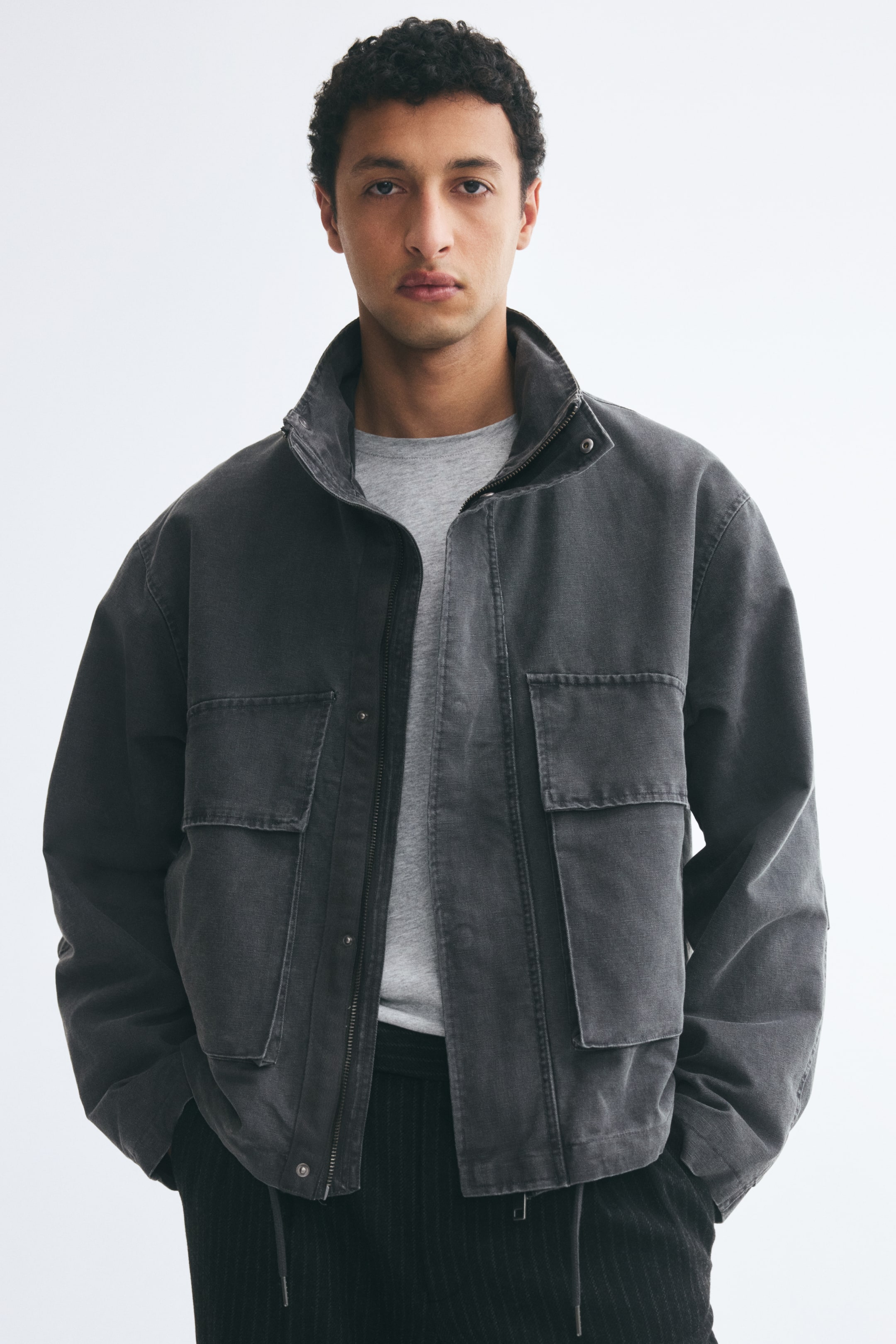 Xem ảnh lớn hơn: Người mẫu đang mặc một chiếc áo khoác vải canvas cotton màu xám đậm bên ngoài áo thun màu xám nhạt và quần tối màu. Áo khoác có khóa kéo phía trước mở kèm nẹp chắn gió, cổ đứng, vai xuôi và các túi hộp lớn có nắp ở mặt trước.