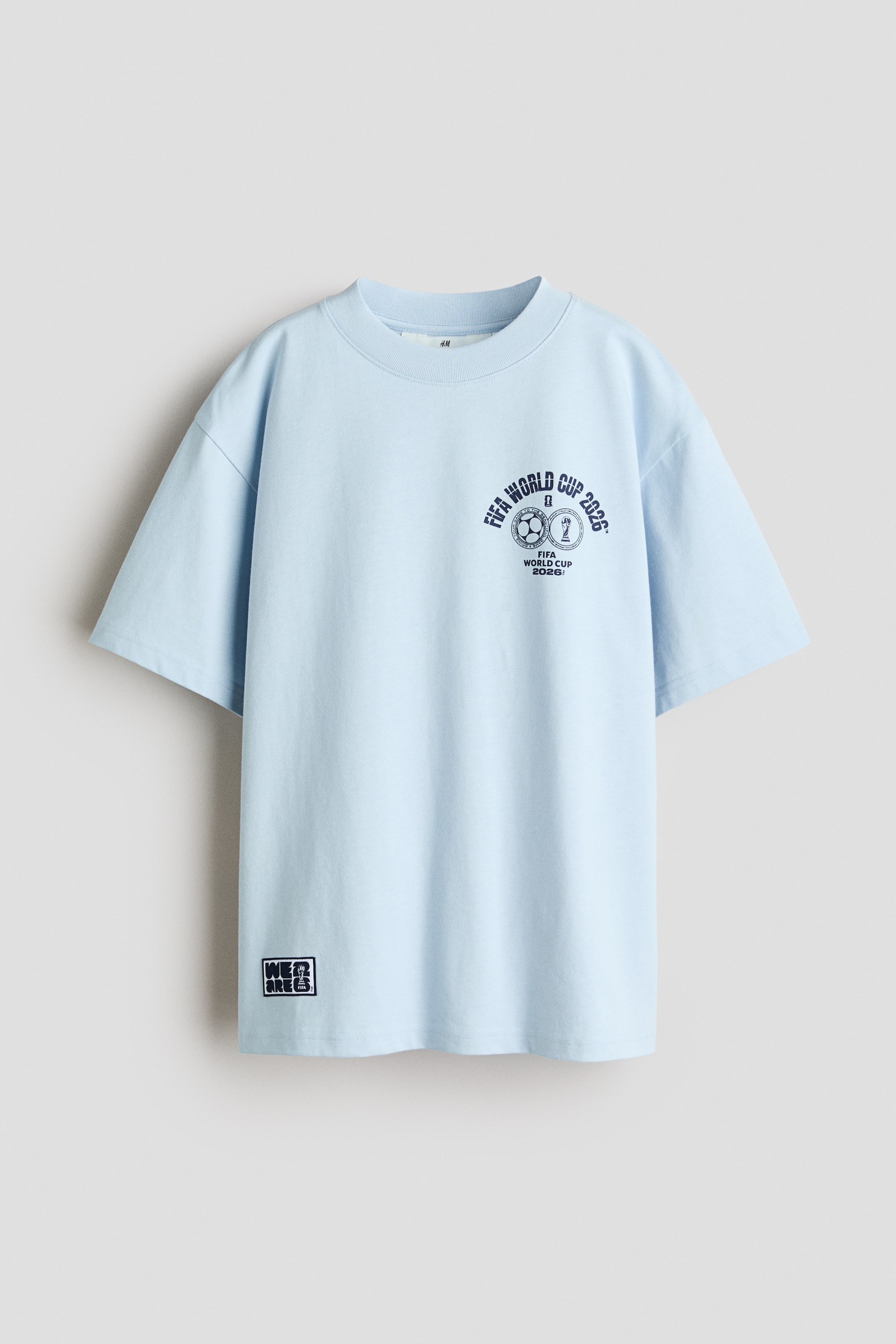 Größeres Bild ansehen: Ein hellblaues Crewneck-T-Shirt zeigt seine Vorderseite mit einem gerippten Halsausschnitt, einer oversized Passform und kurzen Ärmeln. Ein dunkelblauer "FIFA WORLD CUP 2026"-Grafikdruck mit einem Fußball und einer Trophäe ist auf der linken Brust des Trägers zu sehen, während ein "WE ARE"-Grafikdruck am linken unteren Saum des Trägers erscheint.