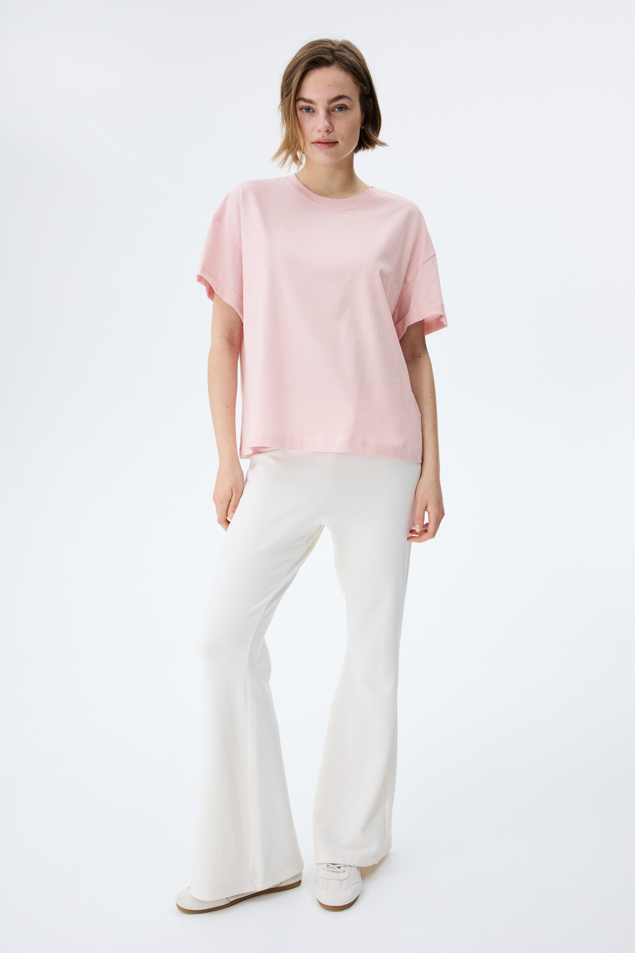 Ingrandisci l'immagine: La t-shirt rosa oversize presenta un girocollo e maniche corte, ed è abbinata a pantaloni bianchi a zampa. I pantaloni mostrano una vestibilità aderente sulla coscia e un'ampia svasatura dal ginocchio.