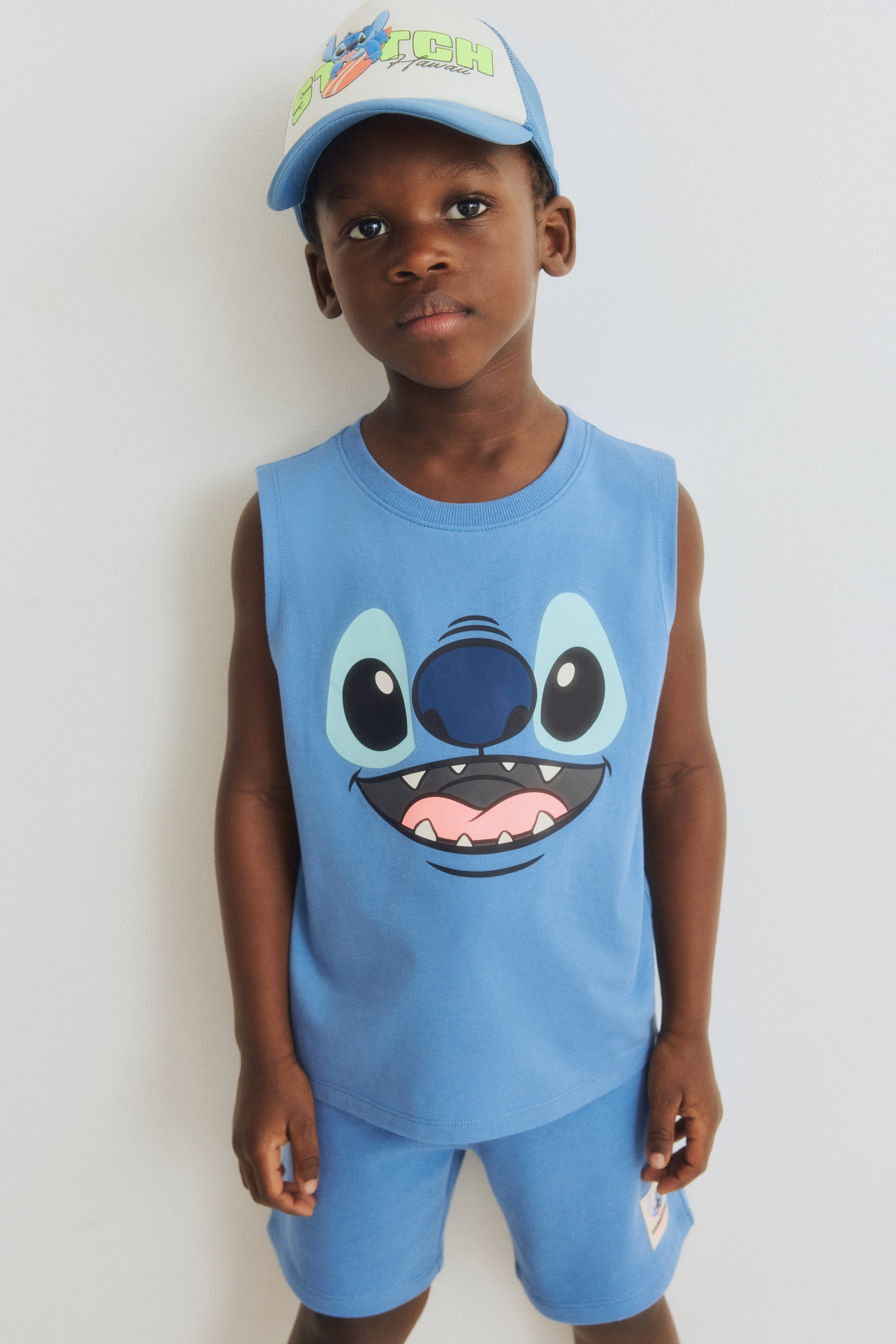 Agrandir l'image: Un haut sans manches bleu clair, avec un grand graphique du visage de Stitch à l'avant, est porté avec un short assorti bleu clair. Une casquette de camionneur bleu clair et blanche, affichant un graphique "Stitch Hawaii" avec Stitch sur une planche de surf, est également présentée.