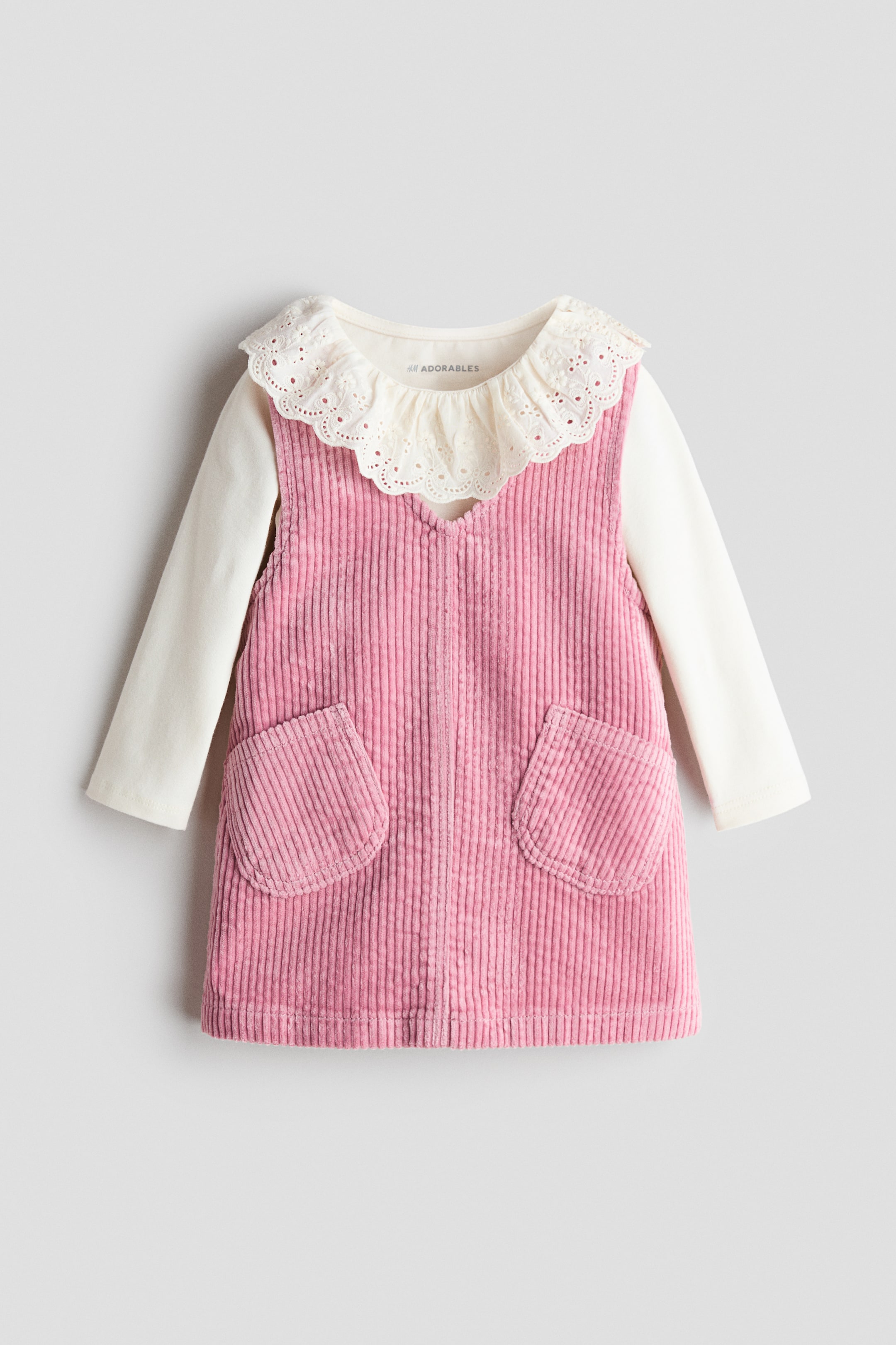 View larger image: The H&M Adorables set showcases a pink corduroy pinafore dress, characterized by a V-shaped neckline, shoulder straps, and two front patch pockets, layered over a cream long-sleeved top. The top features a ruffled, scalloped collar with broderie anglaise detailing, visible beneath the dress's V-neck.