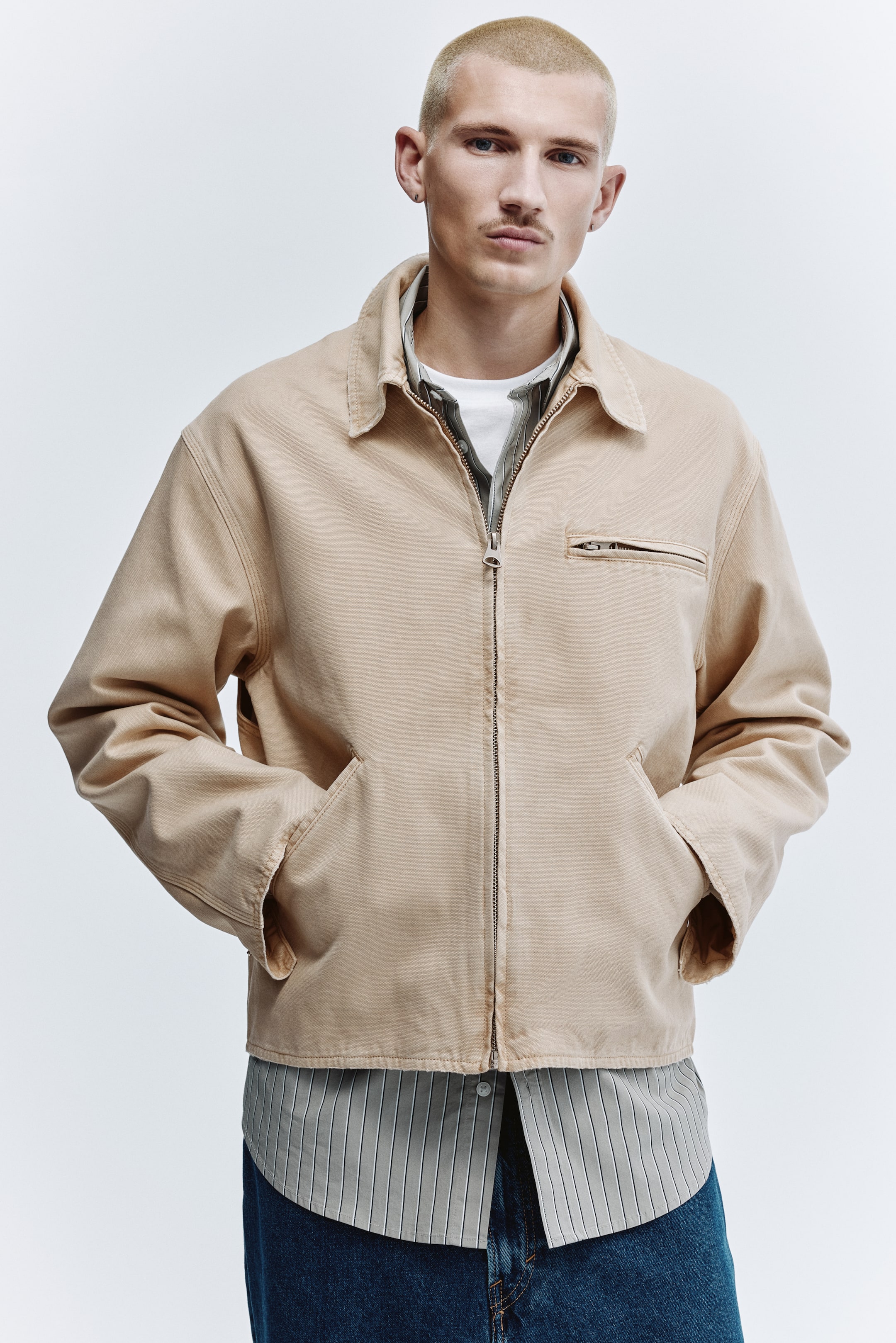 View larger image: A beige workwear jacket faces forward, featuring a full-zip front, a pointed collar, two large patch pockets at the waist, and a zippered chest pocket on the right. The jacket is worn layered over a striped shirt, emphasising its relaxed fit.