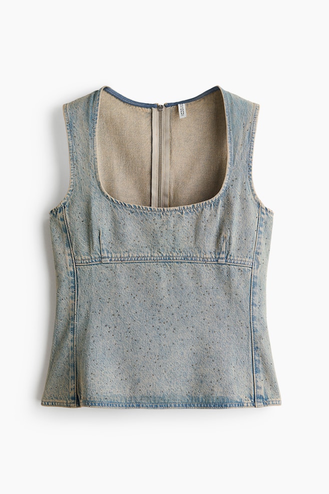 Een mouwloze denim top is van voren afgebeeld en toont een lichtblauwe wassing, een diepe ronde halslijn en overal verspreide rhinestone-versieringen. De top heeft gestructureerde naadpanelen onder de buste en een zichtbare ritssluiting aan de binnenzijde van het achterpand. - 1