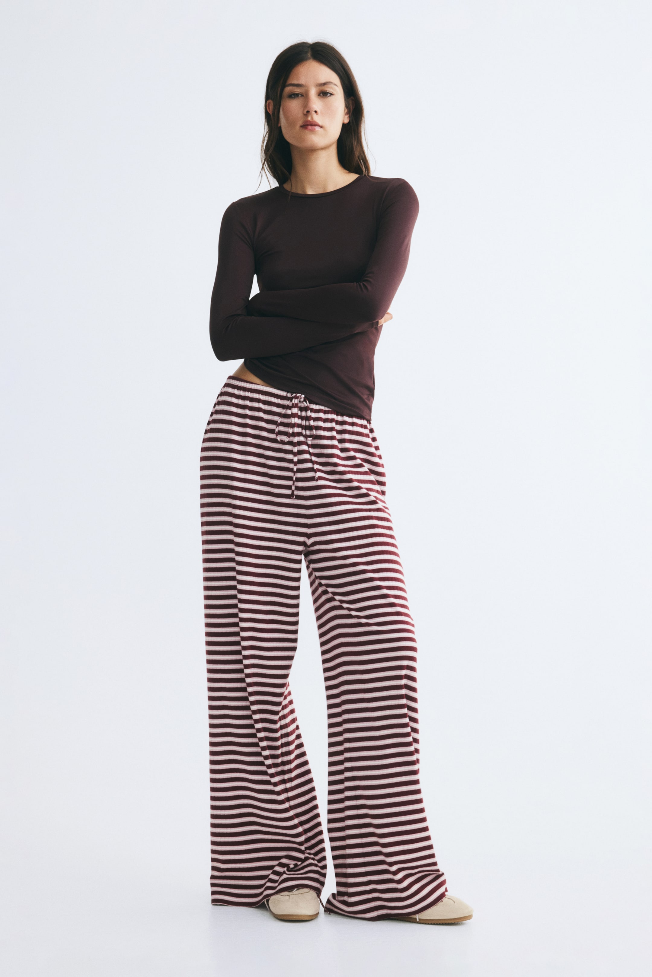 Agrandir l'image: Le mannequin porte un pantalon coupe ample en jersey de coton côtelé, orné de rayures horizontales bordeaux et rose clair. Ce pantalon présente une coupe large et une taille élastiquée avec cordon de serrage à nouer, associé à un haut foncé à manches longues.