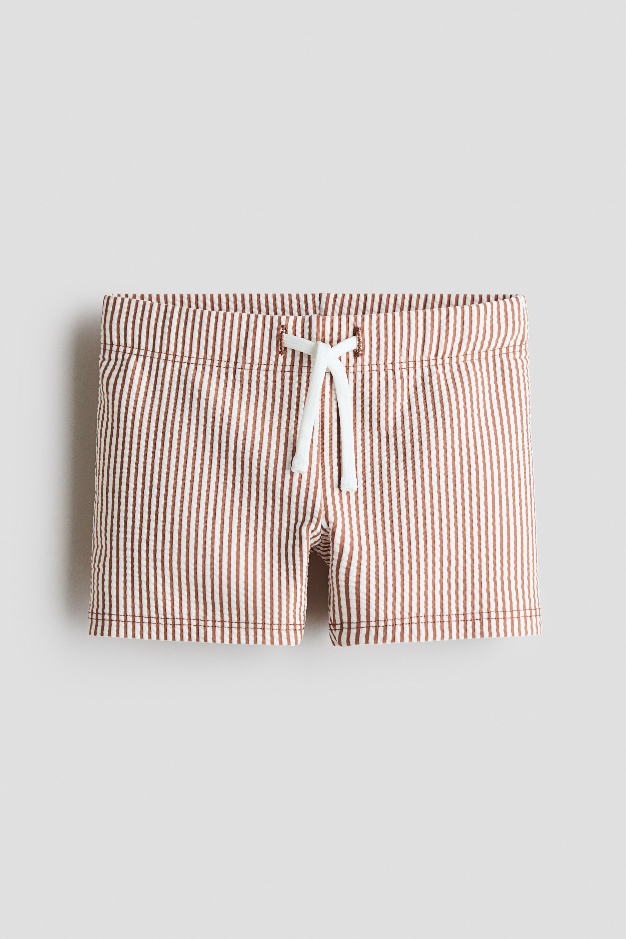 View larger image: These swimming trunks showcase a frontal perspective, adorned with brown and white vertical stripes that boast a seersucker texture. Crafted with an elasticated waistband, they feature a white drawstring at the front, threaded through bronze-toned eyelets, and come fully lined.