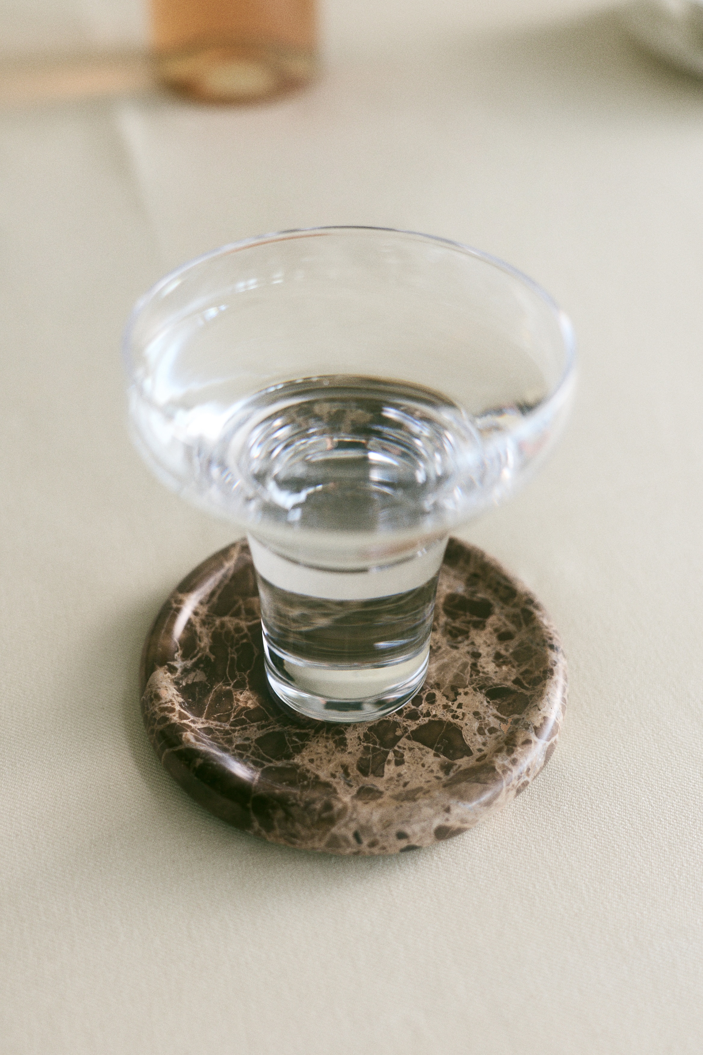 Four round brown marble drink coasters are arranged in a stack, situated next to a light beige rectangular packaging box. The coasters exhibit unique veining patterns, and the box features text on its front that reads "FOR THE PERFECT HOST DRINK COASTERS SET OF 4 brown marble".