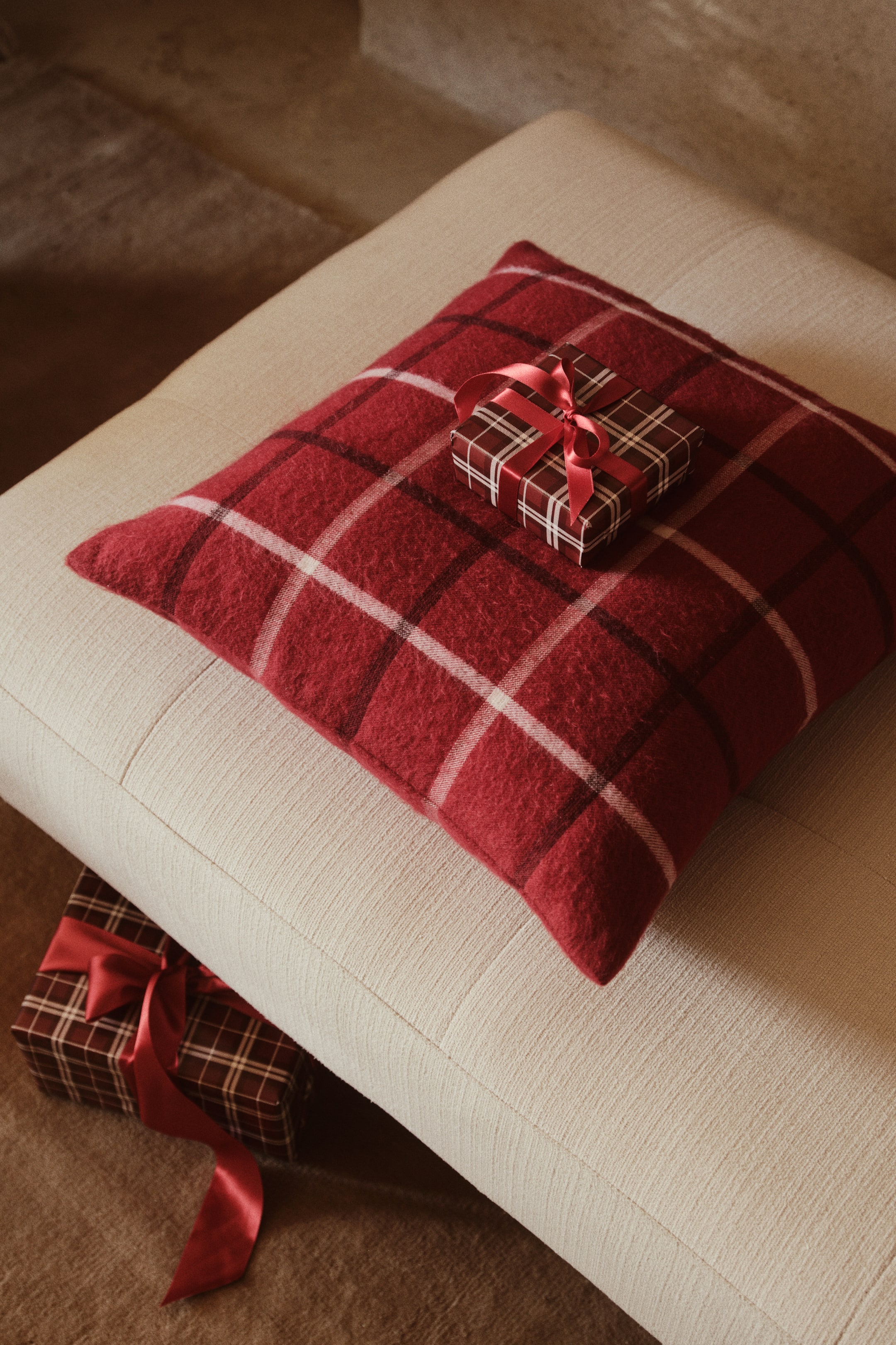 View larger image: A square cushion cover in a red and cream plaid wool-blend jacquard weave with a fuzzy, brushed finish is resting on a light-coloured upholstered bench. A small wrapped gift box is sitting on top of the cushion, showcasing its soft and thick texture.