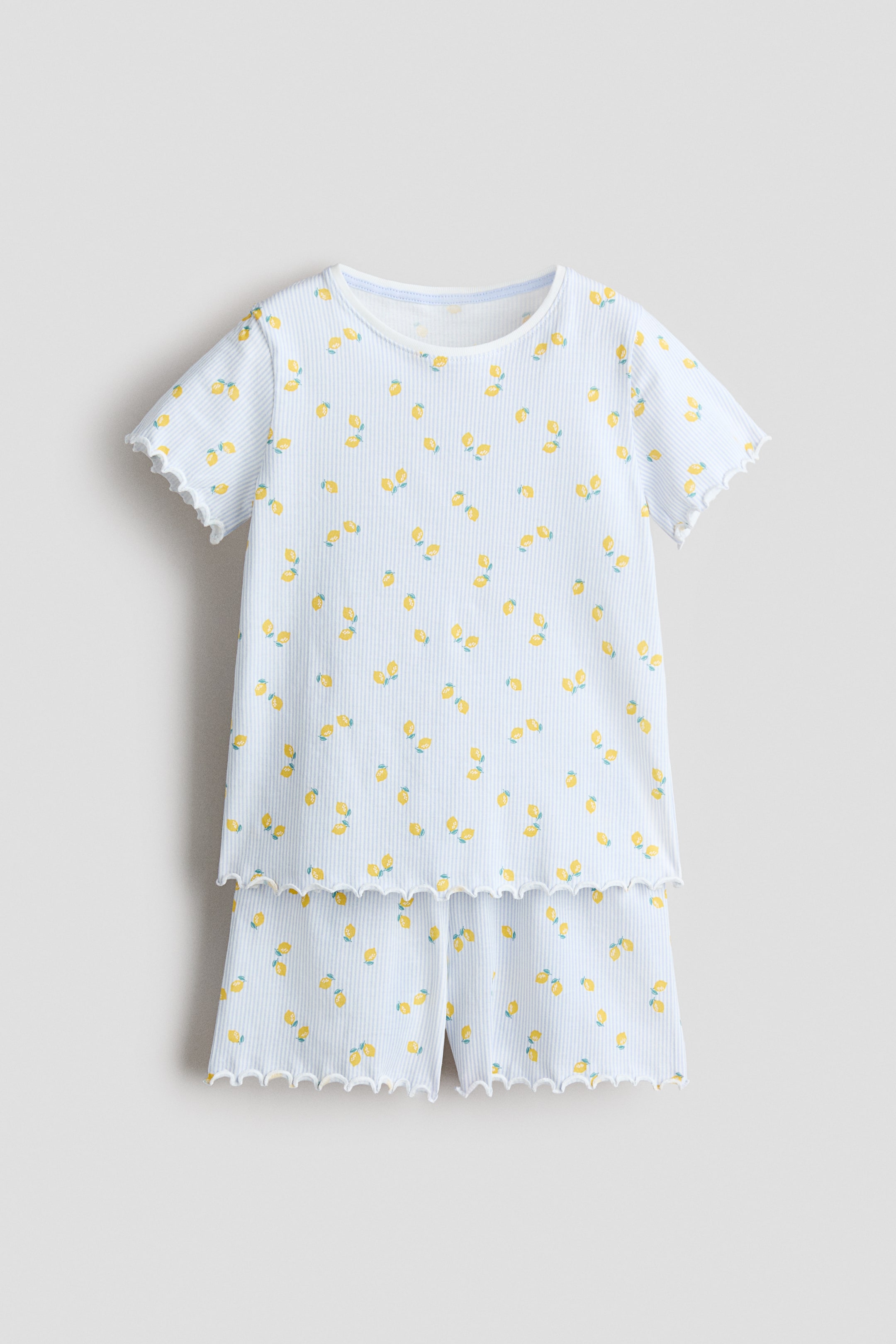 View larger image: A two-piece pajama set featuring a short-sleeved top and shorts, showcased from the front. The light blue ribbed fabric displays thin white vertical stripes and a scattered pattern of small yellow lemons with green leaves. Both the top's rounded neckline, short sleeves, and hem, along with the shorts' leg openings, are finished with a white scalloped edge.