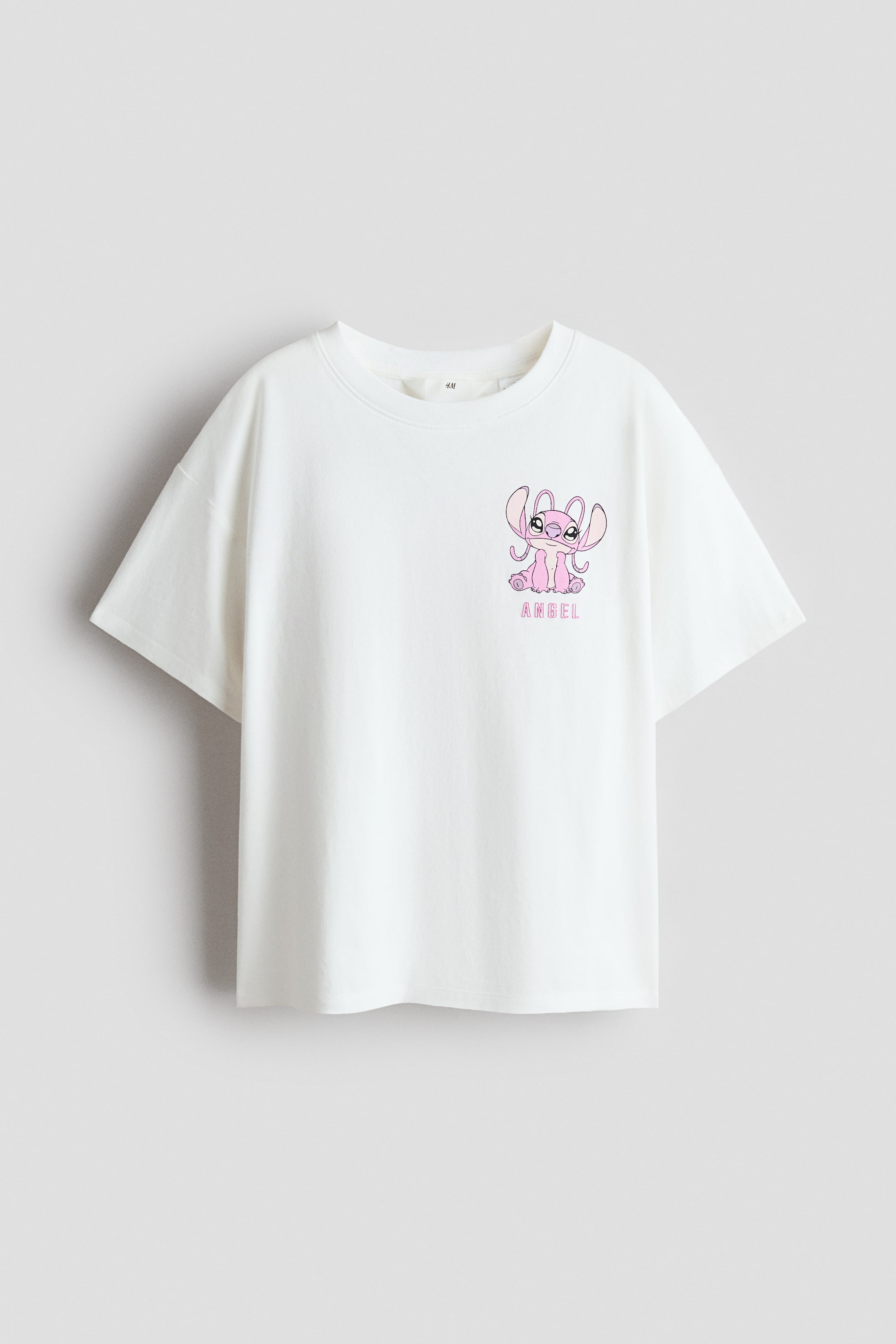 查看大圖: 一件柔軟白色棉質針織布的大號T恤正面展示。它的特色是圓形羅紋飾邊領口和落肩設計,左胸印有粉紅色卡通角色以及"ANGEL"字樣。