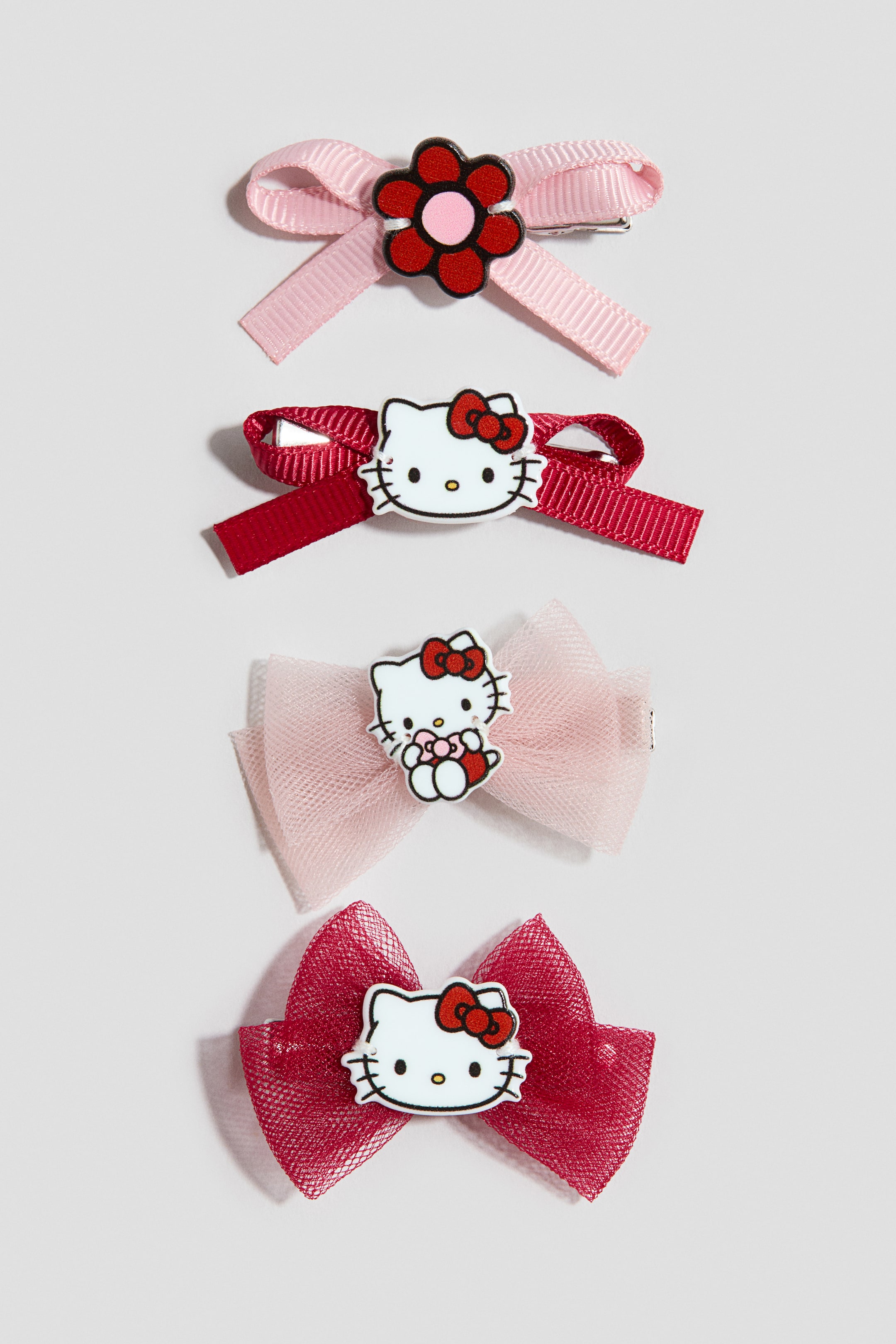 View larger image: A set of four decorative hair clips is shown front-facing in a vertical arrangement, showcasing various bow styles and charms. The top pair features grosgrain ribbon bows in light pink with a flower charm and dark red with a Hello Kitty face, while the bottom pair features light pink and dark red tulle bows accented with Hello Kitty charms.