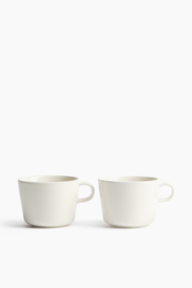 Two off-white ceramic cups are standing side-by-side, facing front. Each cup features a subtly tapered body and a C-shaped handle. - 1