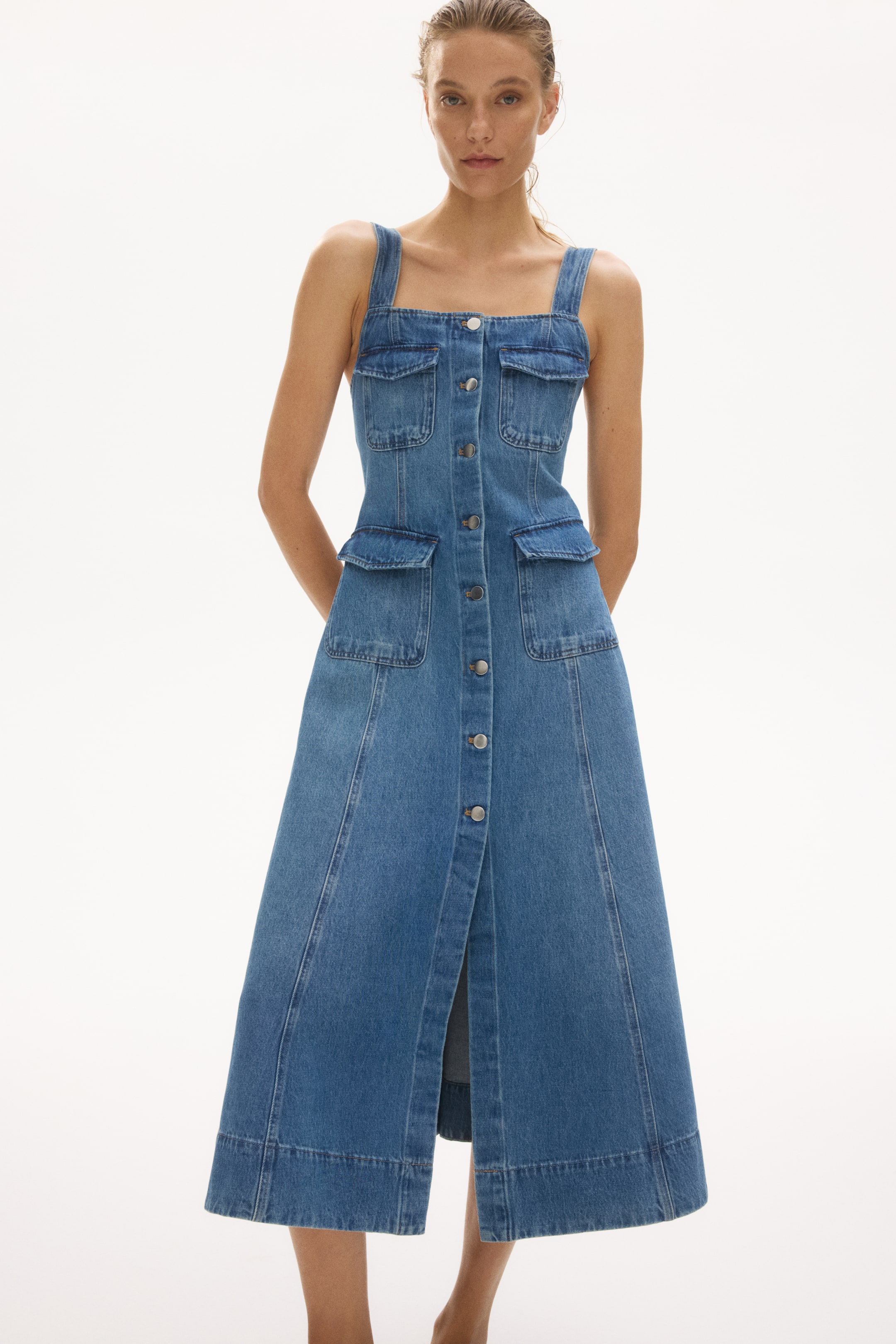 Ampliar la imagen: Este vestido de mezclilla a media pierna presenta un escote cuadrado, tirantes anchos y botones que recorren su parte frontal. También cuenta con bolsillos de solapa en el pecho, bolsillos de solapa frontales, un corpiño ajustado, una falda acampanada y costuras visibles en la parte delantera.