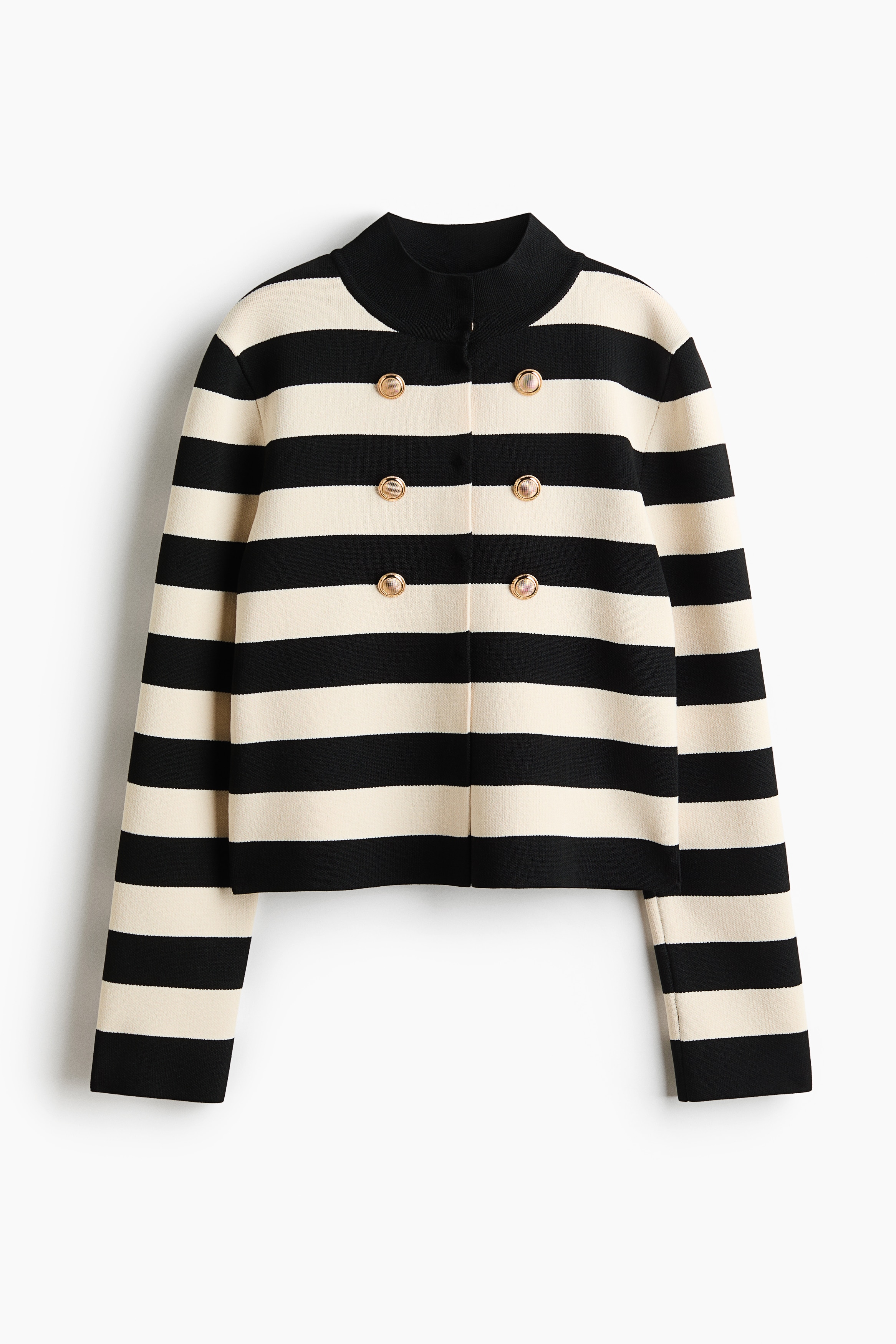 A striped black and cream knit cardigan is being worn by the model, featuring a black ribbed stand collar, dropped shoulders, and decorative gold-tone buttons on the front. The model is sitting with folded arms, styling the cardigan with dark denim jeans.