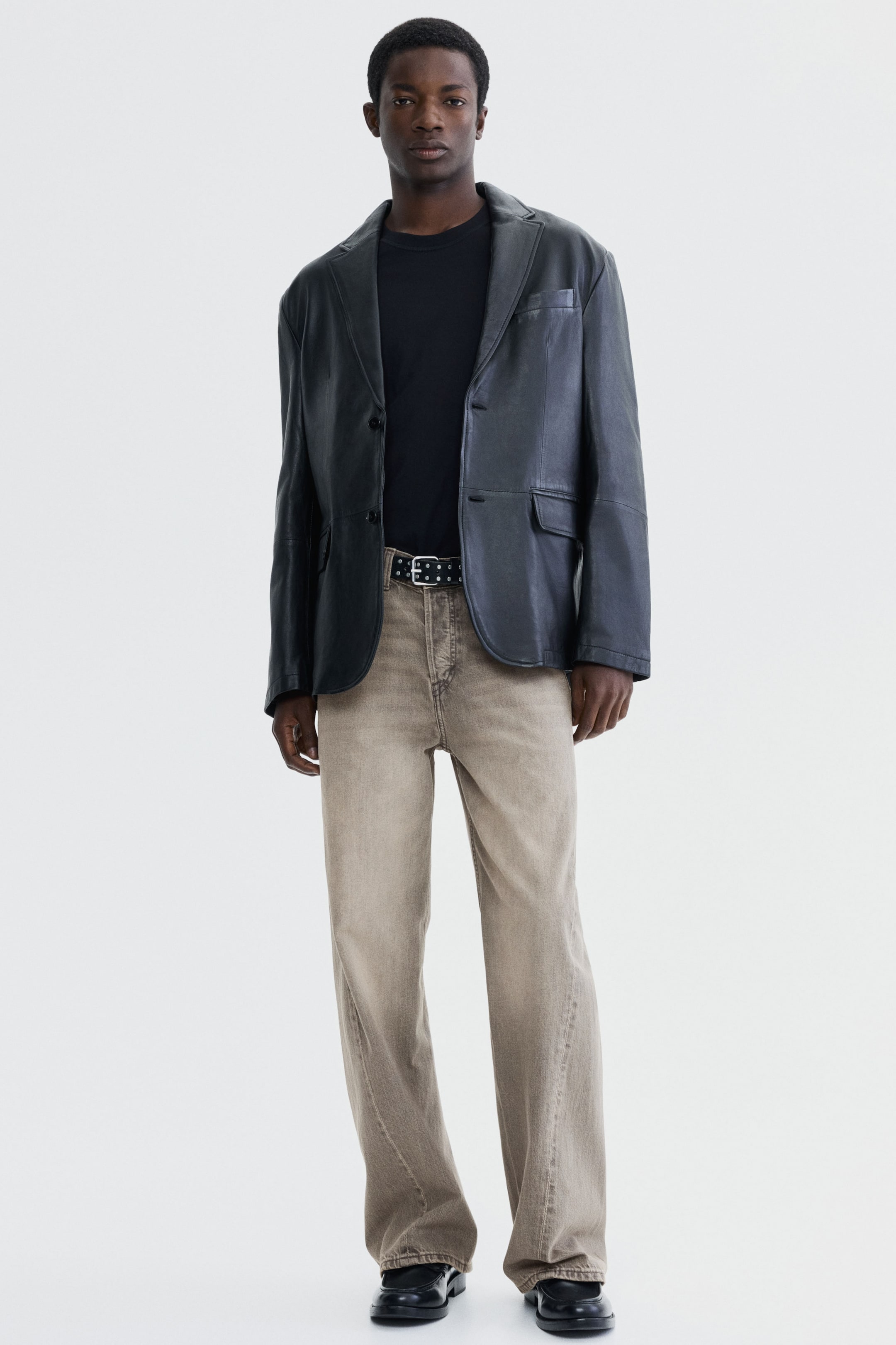 Se større billede: Afslappede, greige jeans med lige ben og fuld længde, der samler sig let ved anklerne. Et nittet sort bælte strammer taljen ind.