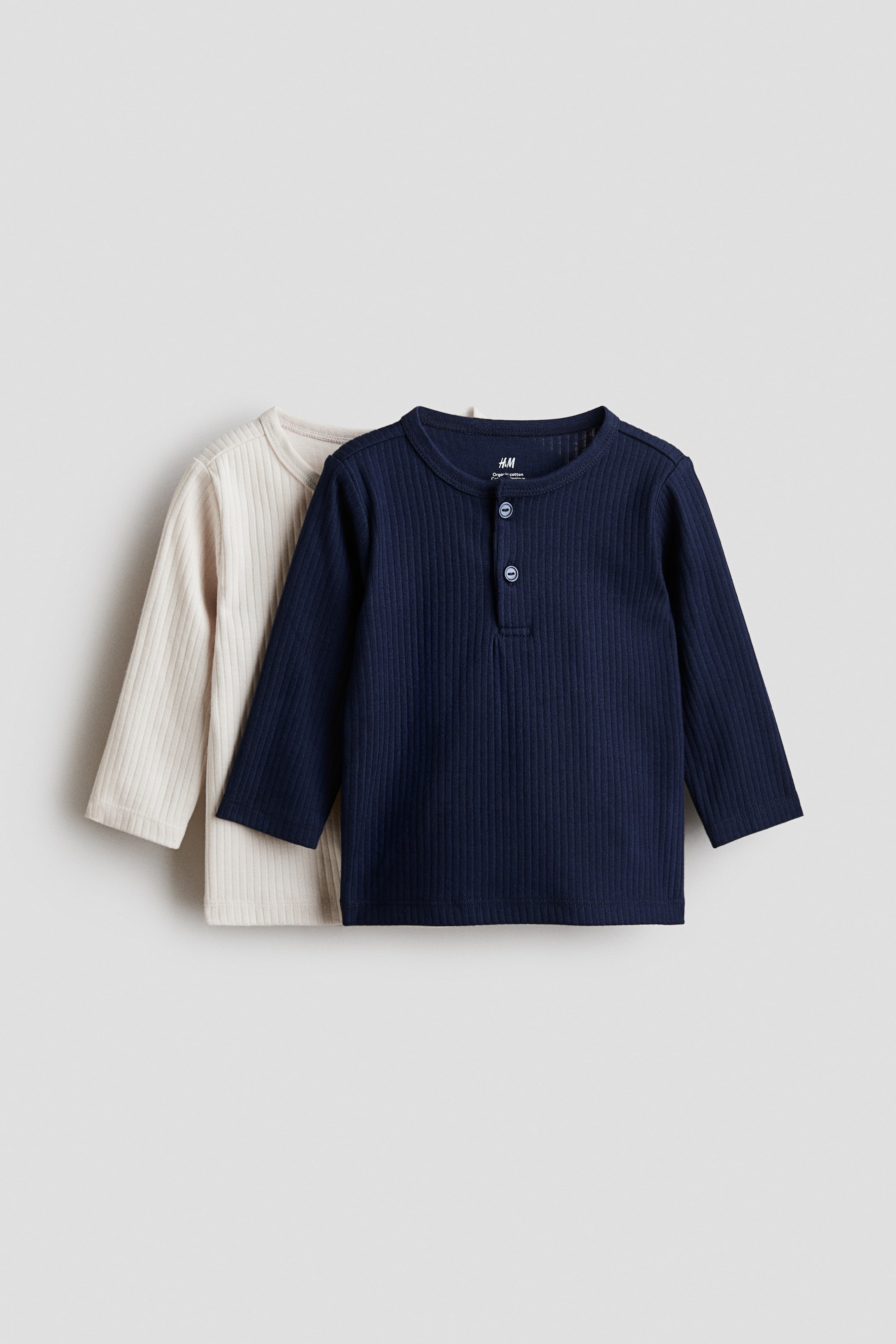 View larger image: Two long-sleeved ribbed shirts are presented from the front. The navy-blue shirt features a round neckline and a Henley placket with three light-coloured buttons, while a light beige shirt is partially visible behind it.