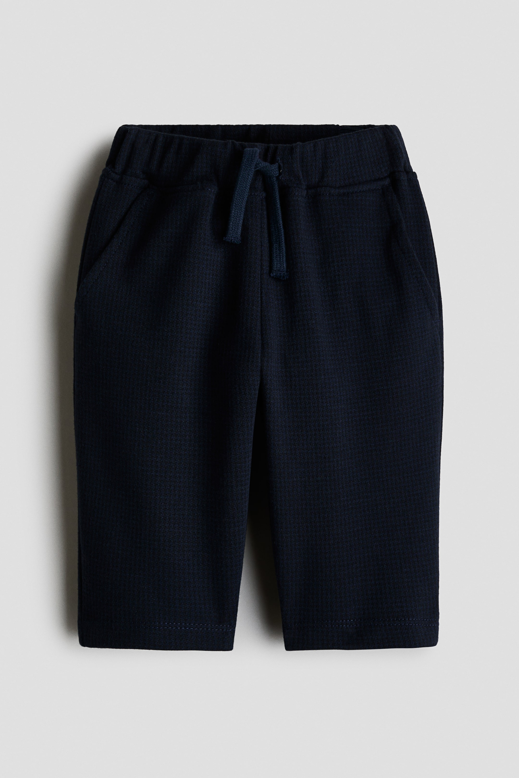 View larger image: A front view of dark navy blue trousers featuring an elasticated waistband with a matching drawstring tied at the centre. The trousers showcase a textured knit fabric, side slant pockets, and straight-cut legs.